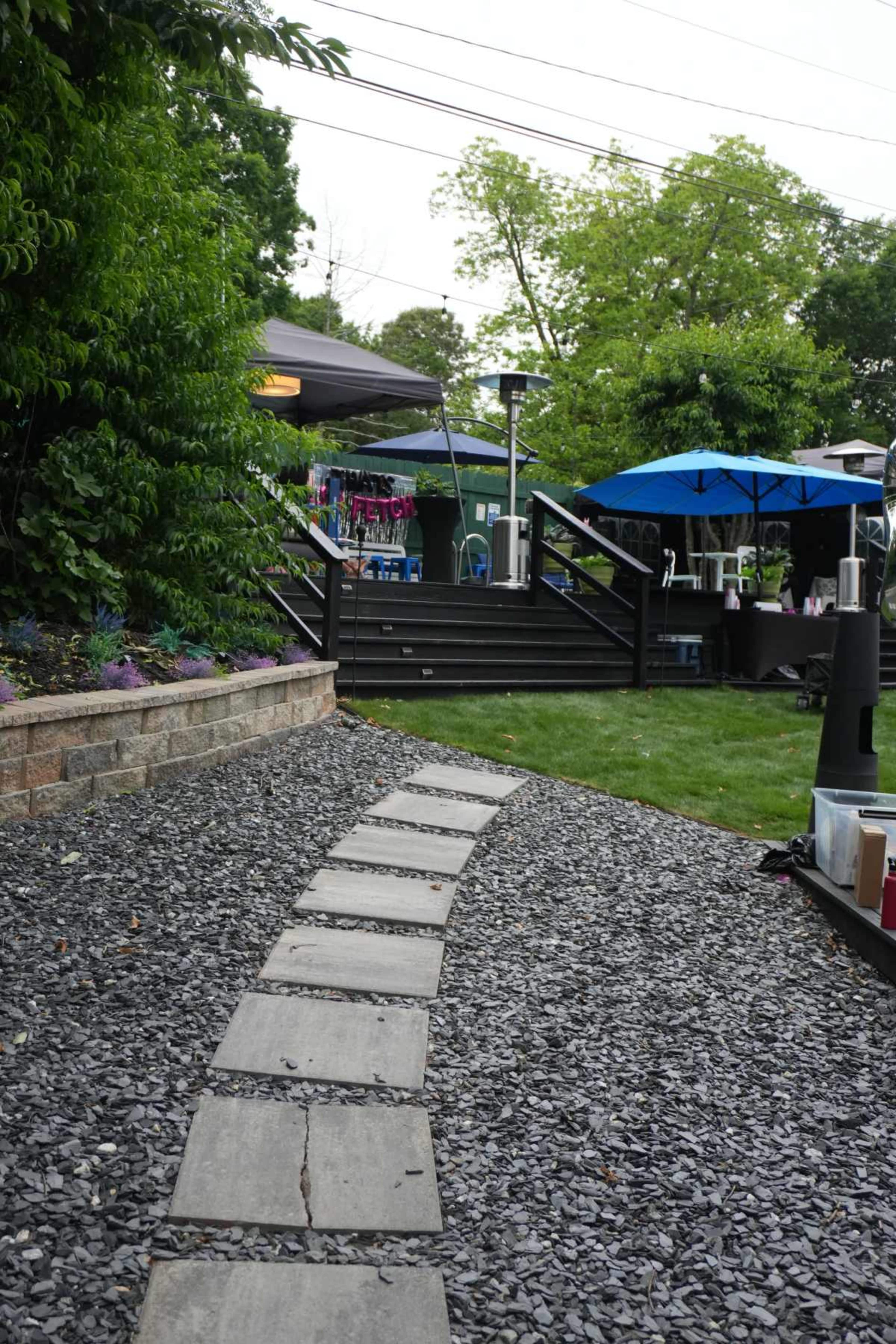 The image shows a curved stone pathway leading through gravel towards a deck area with tents and blue umbrellas.