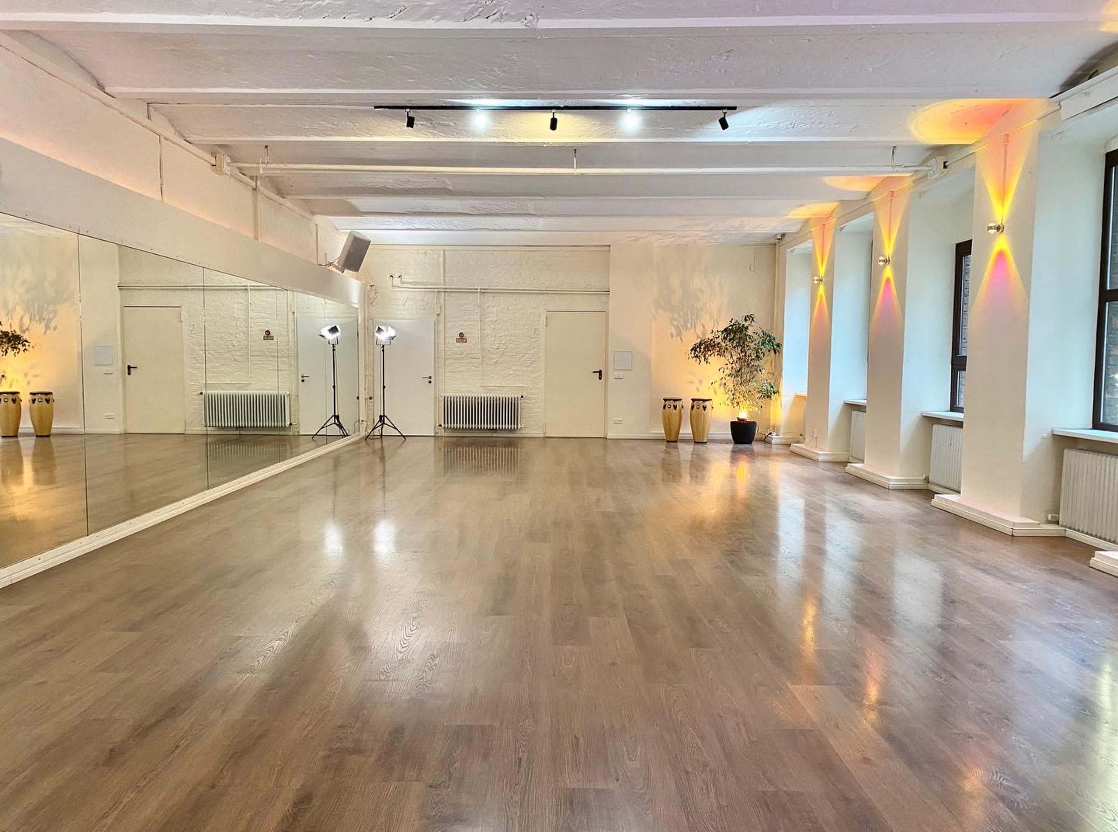 A spacious, well-lit dance studio features wooden flooring, mirrors on one wall, and soft lighting along the other walls.