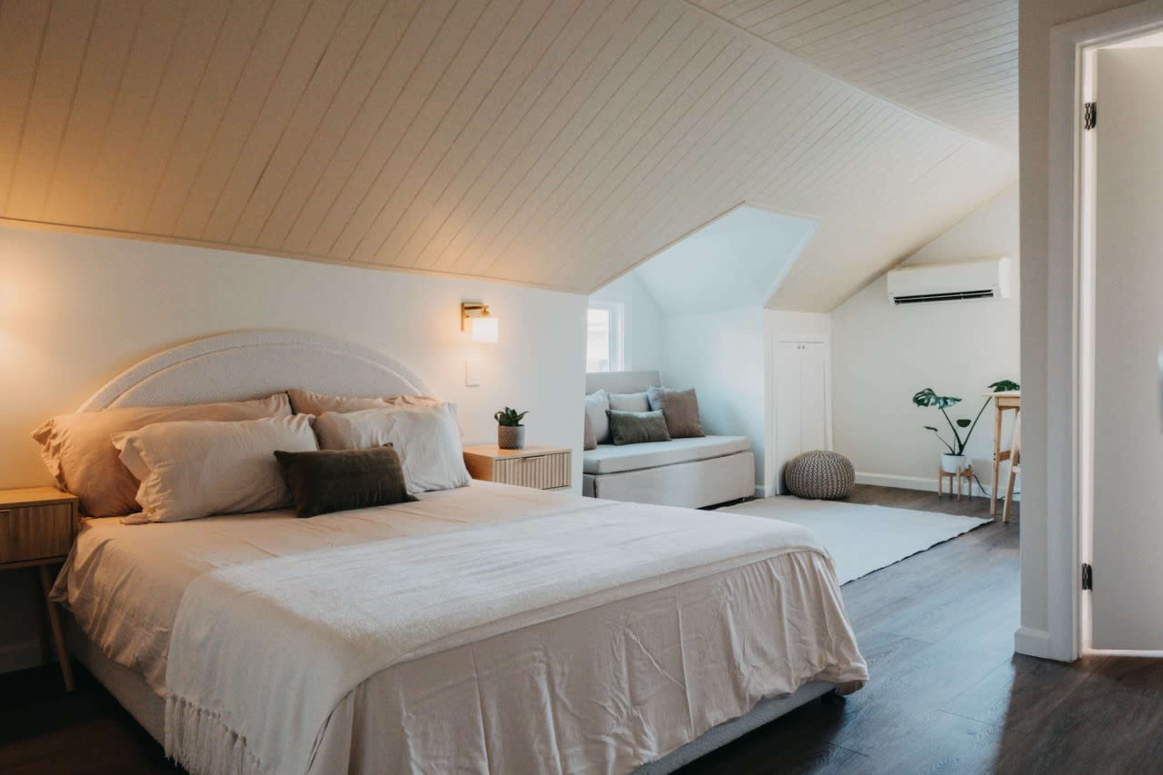 The image shows a modern bedroom with a sloped ceiling, featuring a large bed, a light-colored bedspread, and a small seating area with a couch and decorative plants.
