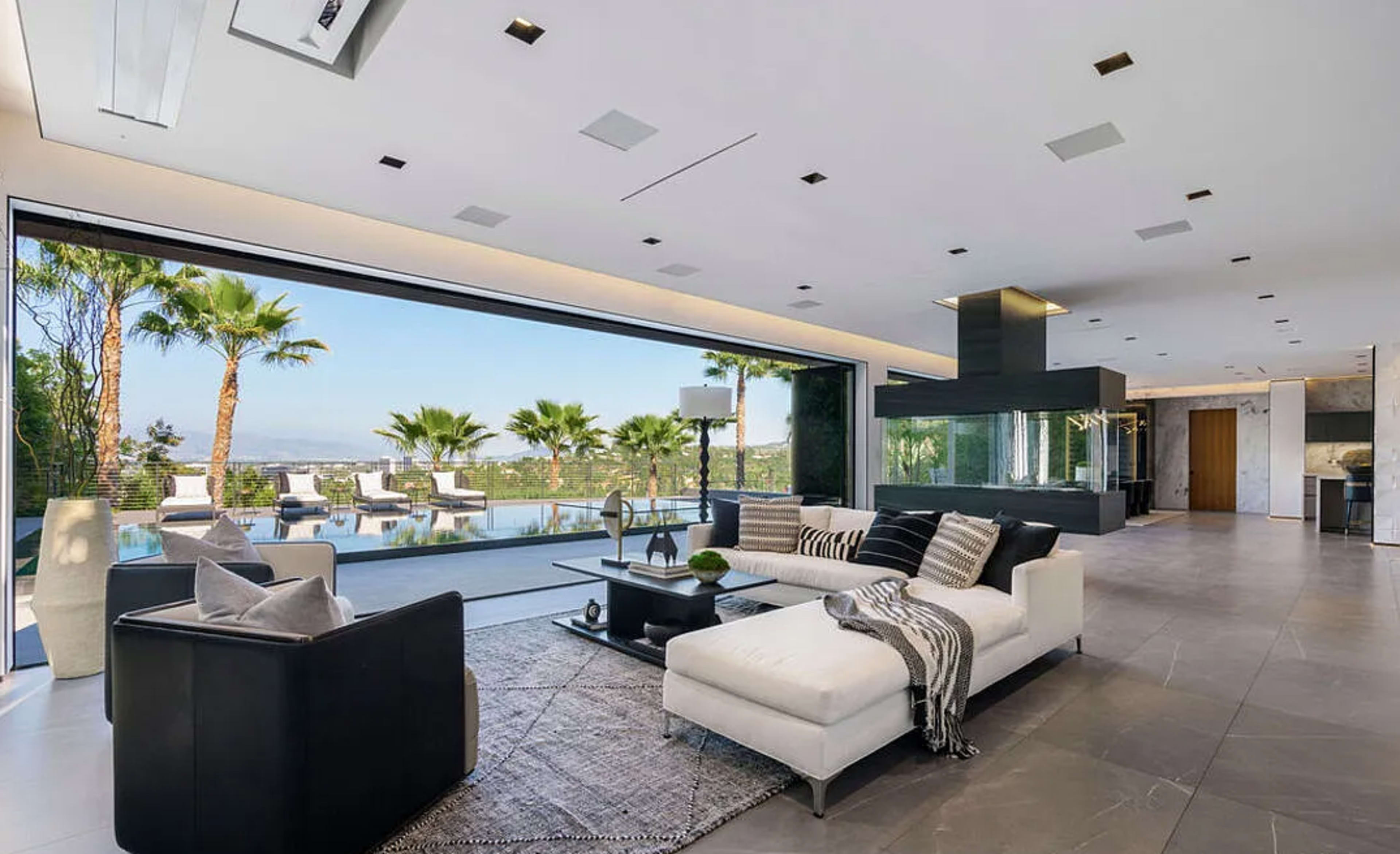The image shows a modern living room with a large window overlooking a pool area surrounded by palm trees.