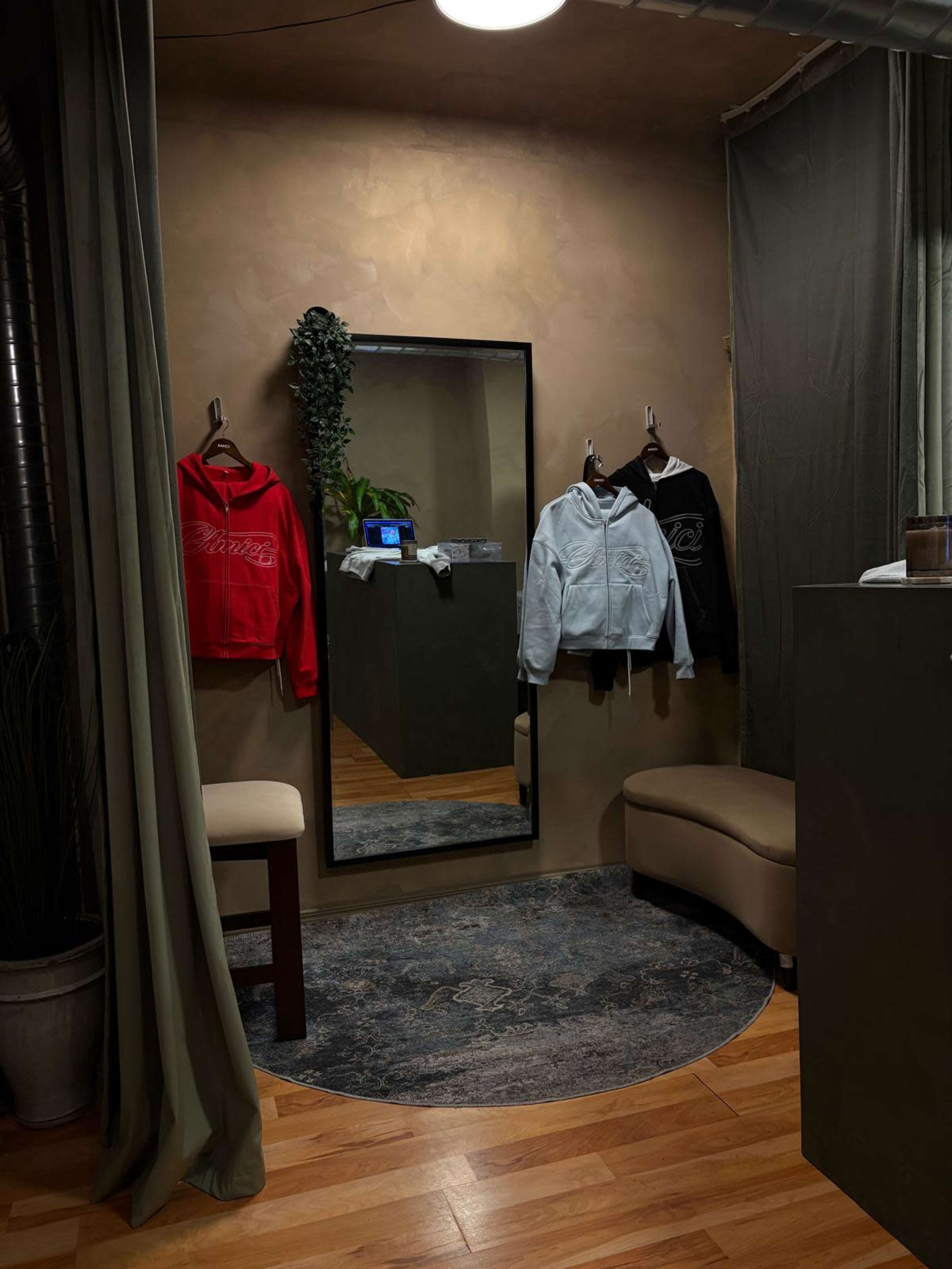 The image shows a clothing fitting area with three hoodies displayed on hangers near a full-length mirror, accompanied by a small seating area and soft lighting.