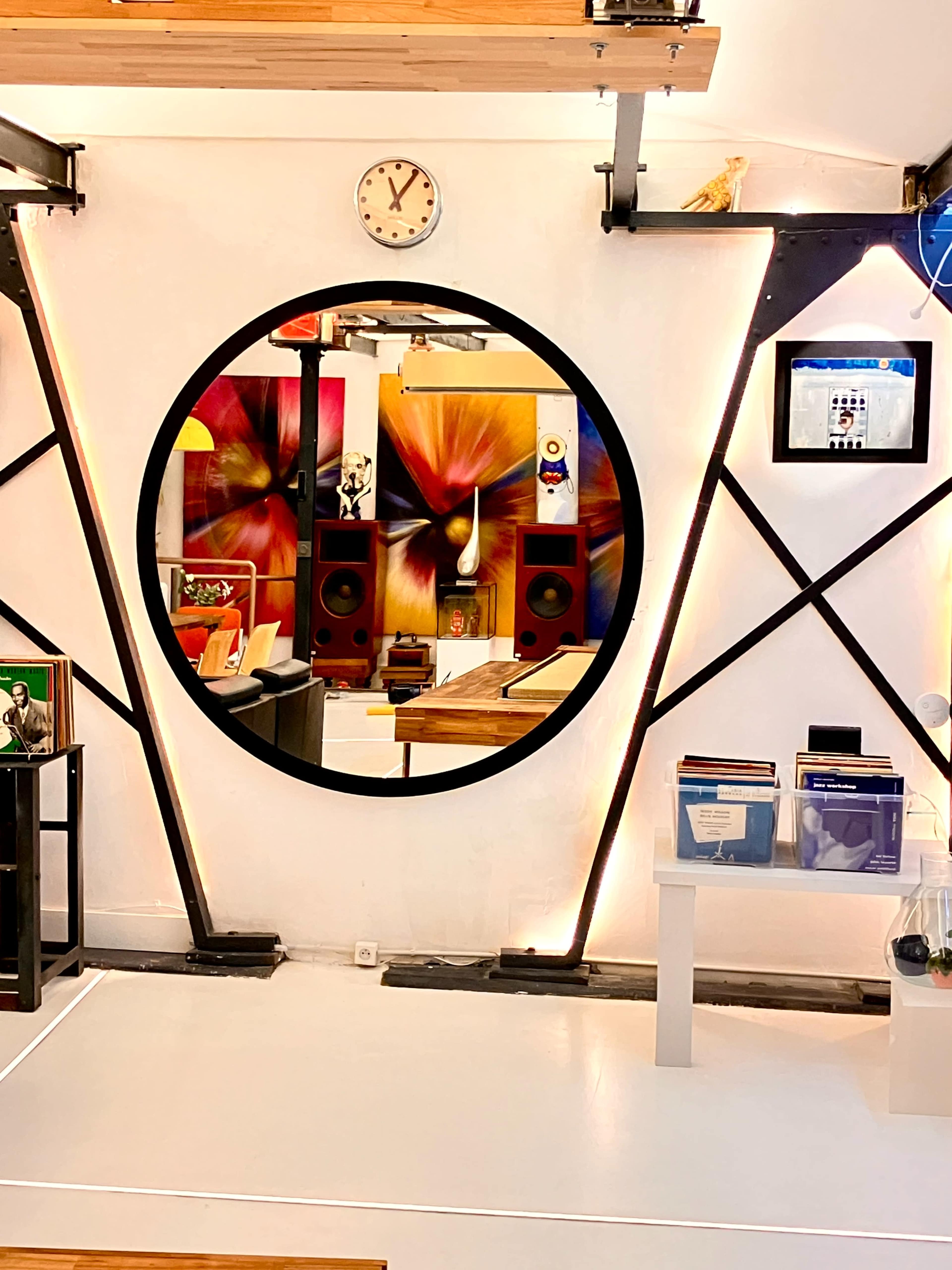 The image features a circular mirror mounted on a wall with a clock above it, framed by angular black supports, while colorful artwork and audio equipment are visible in the background.
