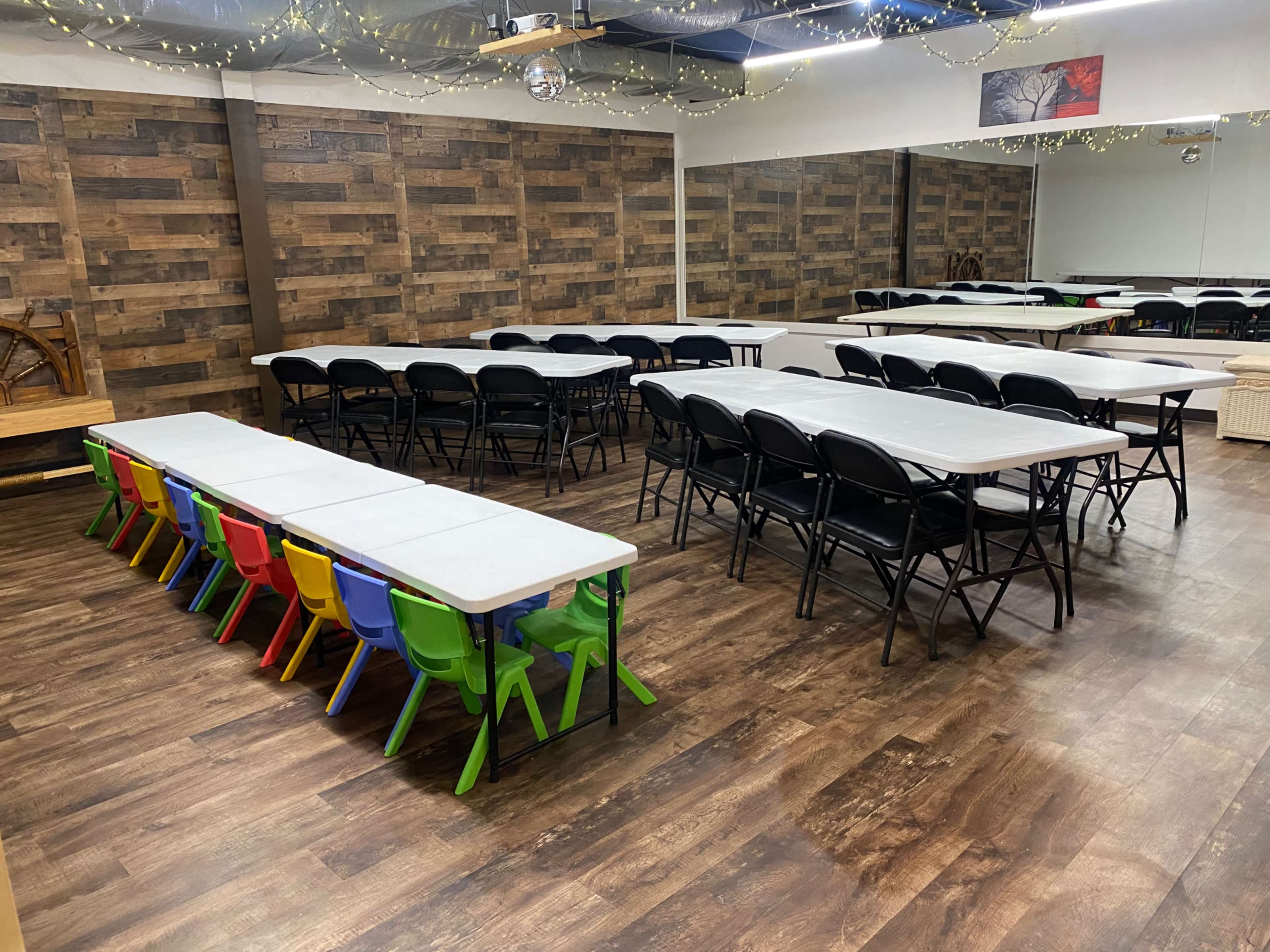 A spacious room features several rows of folding tables and chairs, with colorful plastic chairs lined up at one table against a wooden wall.