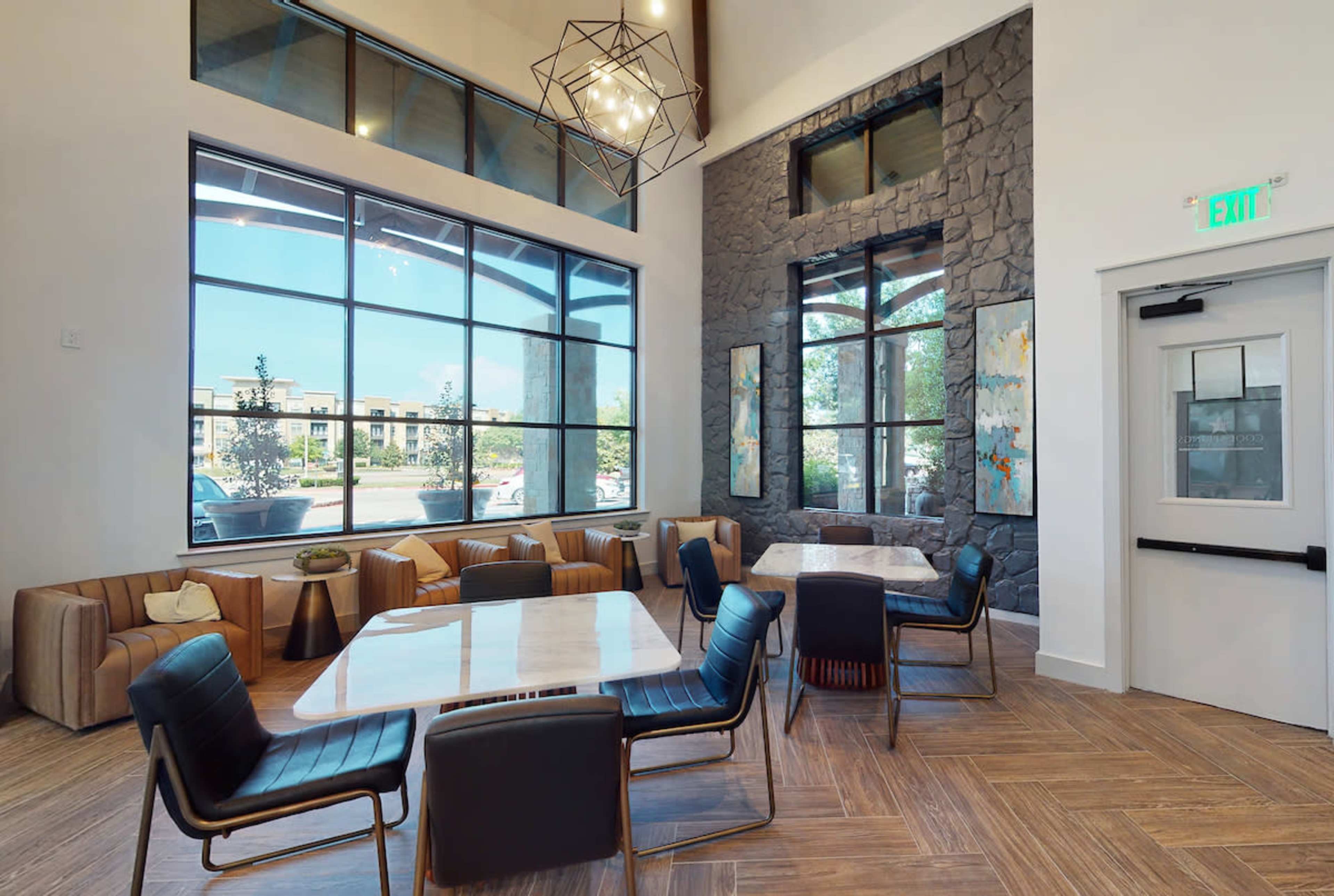 A modern lounge area features large windows, a stone accent wall, and a mix of seating options, including sofas and chairs around wooden tables.