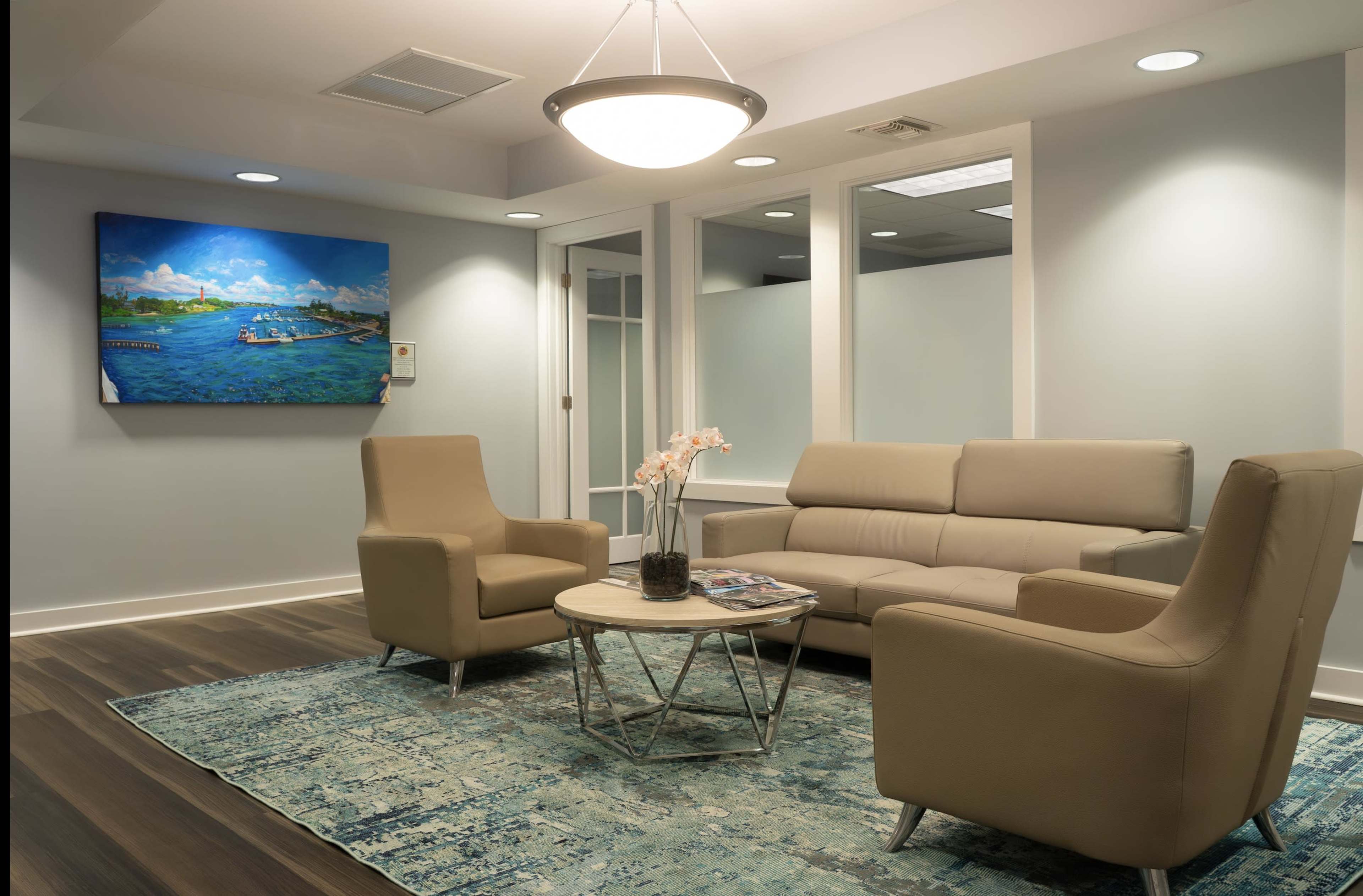The image shows a modern waiting room with two armchairs and a sofa arranged around a round coffee table on a patterned rug, featuring a colorful landscape painting on the wall.