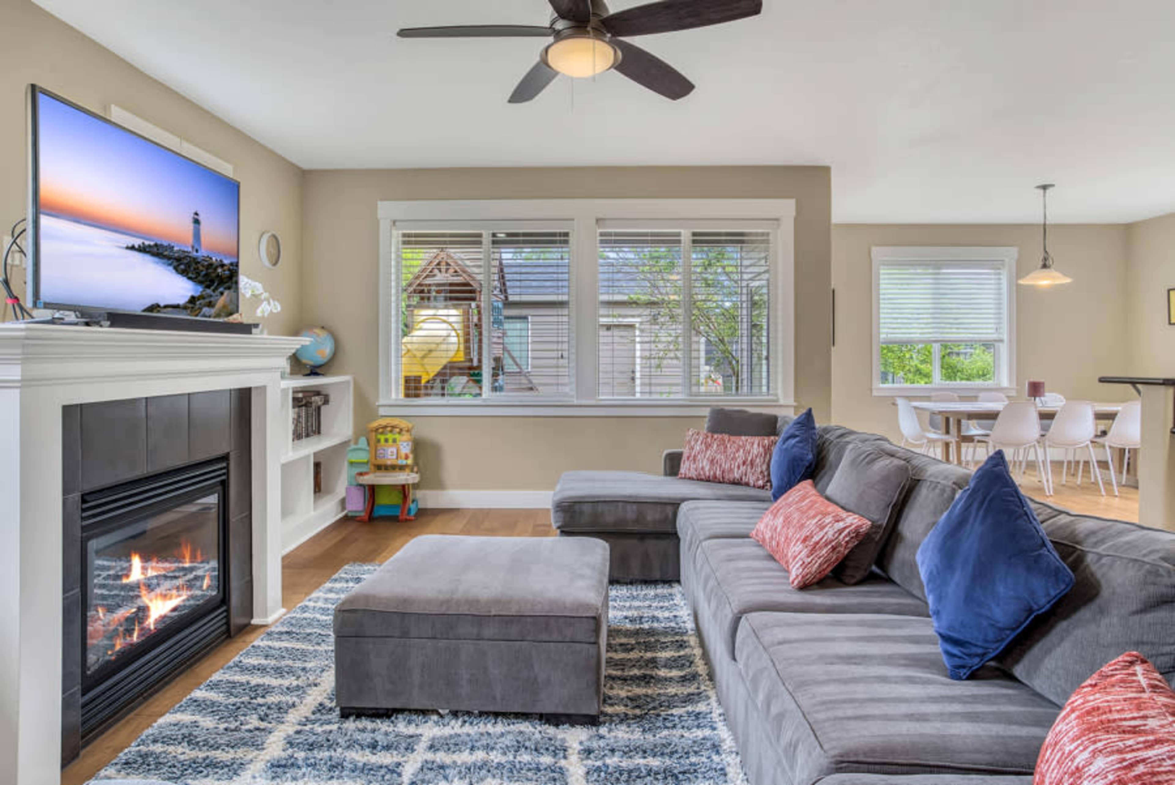 The living room features a gray sectional sofa, a television mounted above a fireplace, and a dining area with modern chairs and a visible play structure outside.