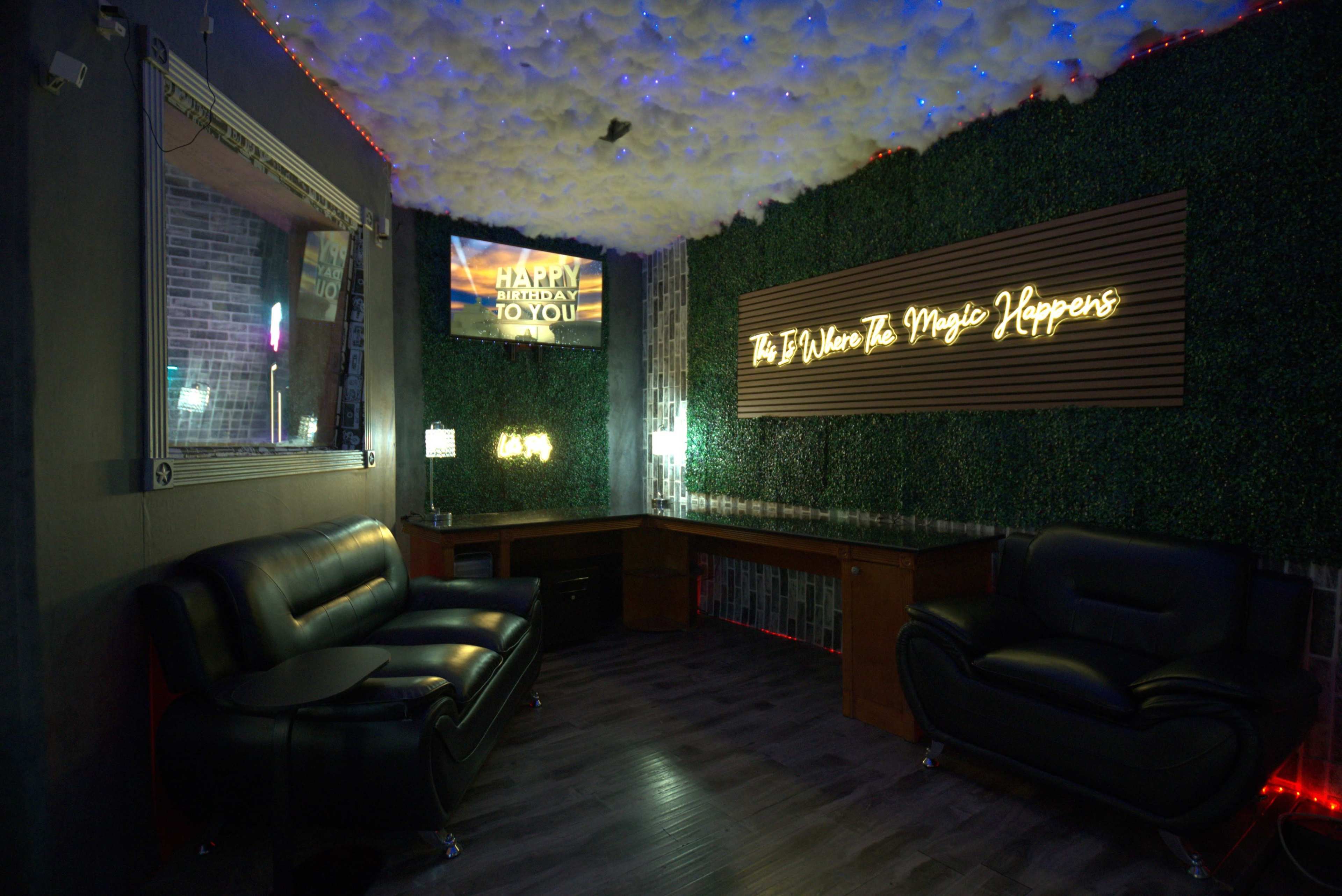 The image shows a dimly lit lounge area with green wall decor, two black leather sofas, and a screen displaying a birthday message.