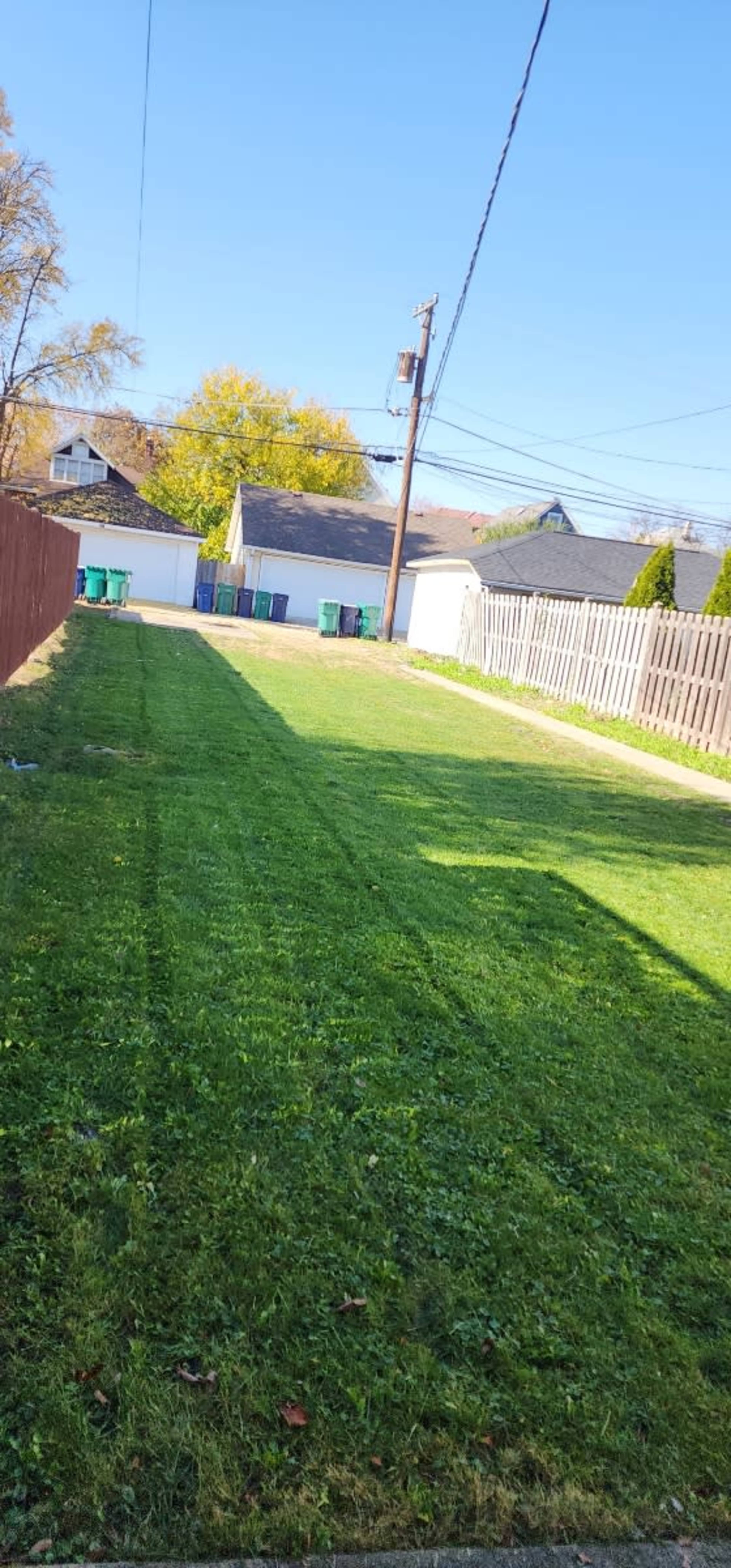 A neatly mowed lawn with green grass, leading to a parking area behind a row of trash bins and a garage under a clear blue sky.