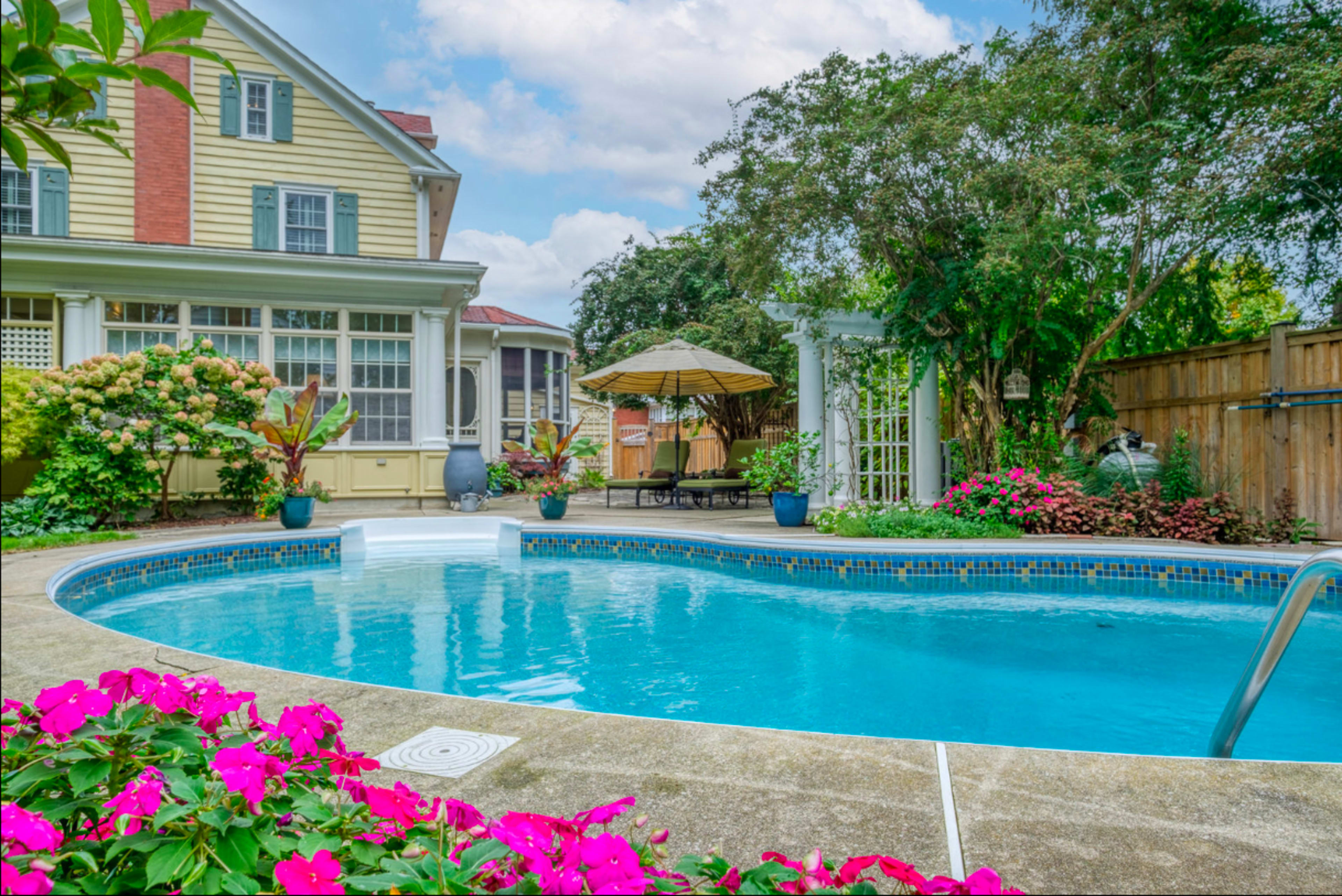 A clear swimming pool surrounded by colorful flowers and lush greenery, with a house and patio area in the background.