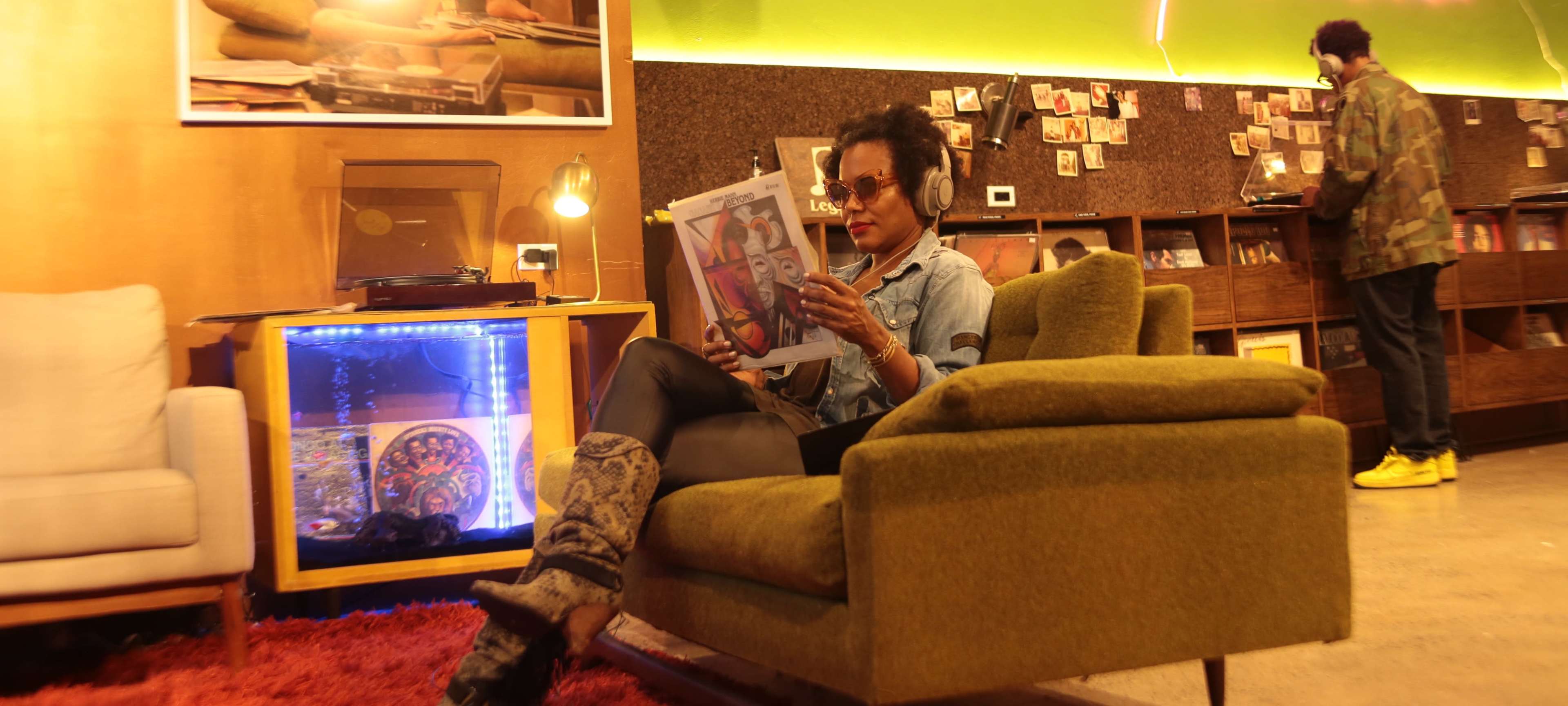 A woman sits on a green chair reading a magazine in a cozy, retro-styled café with wooden decor and a record player nearby.