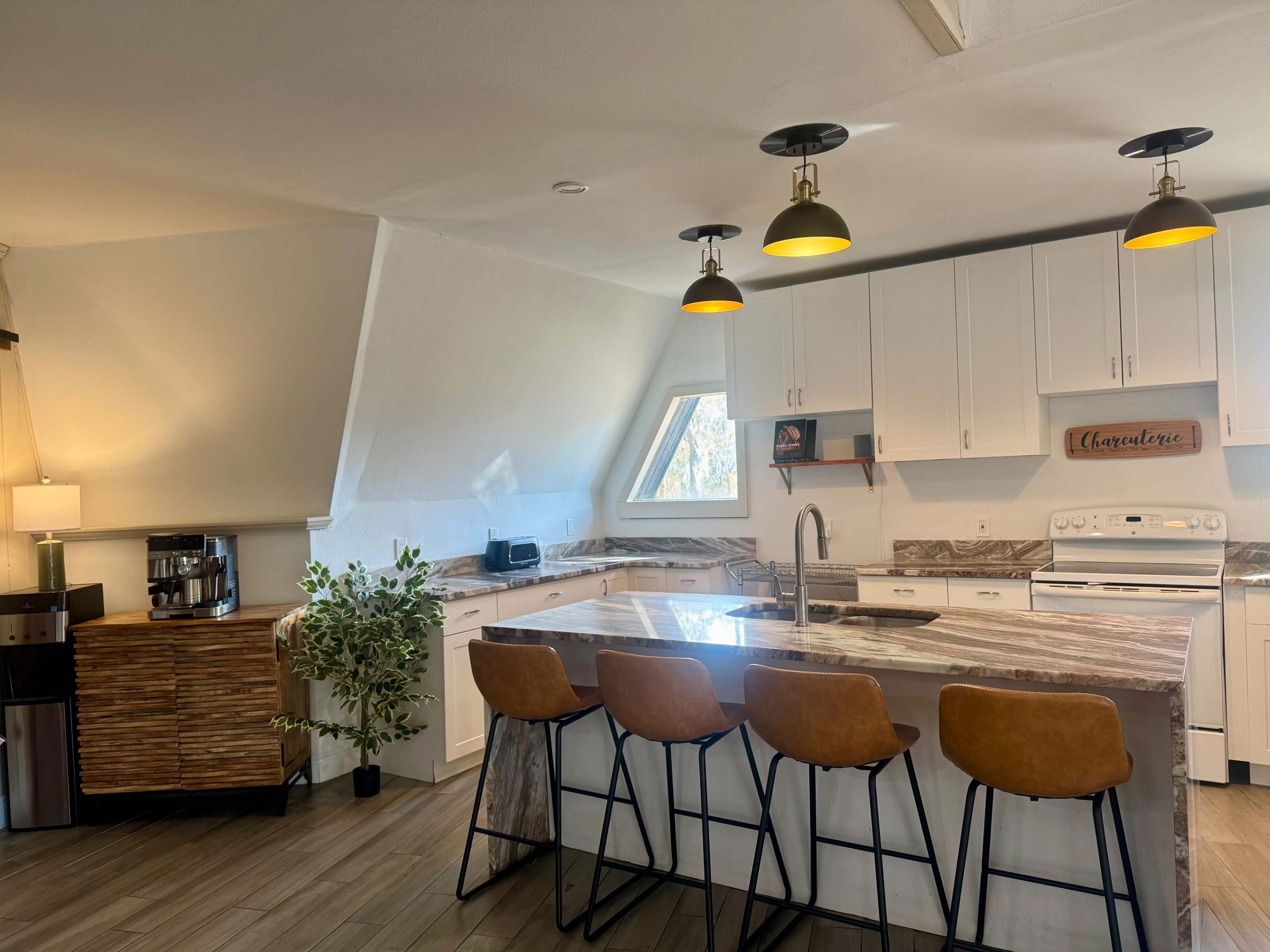 A modern kitchen features a large island with four brown bar stools, sleek white cabinetry, and pendant lighting over the countertop.