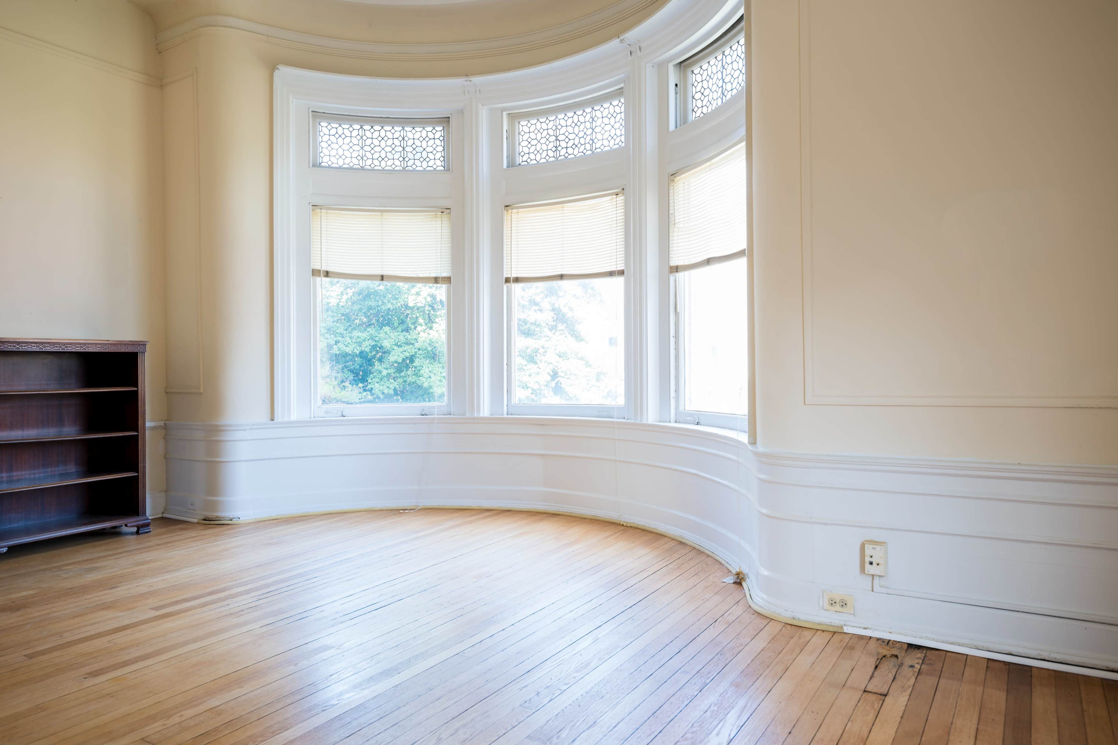 The image shows an empty room with wooden flooring, a curved wall of windows, and a bookshelf along one side.