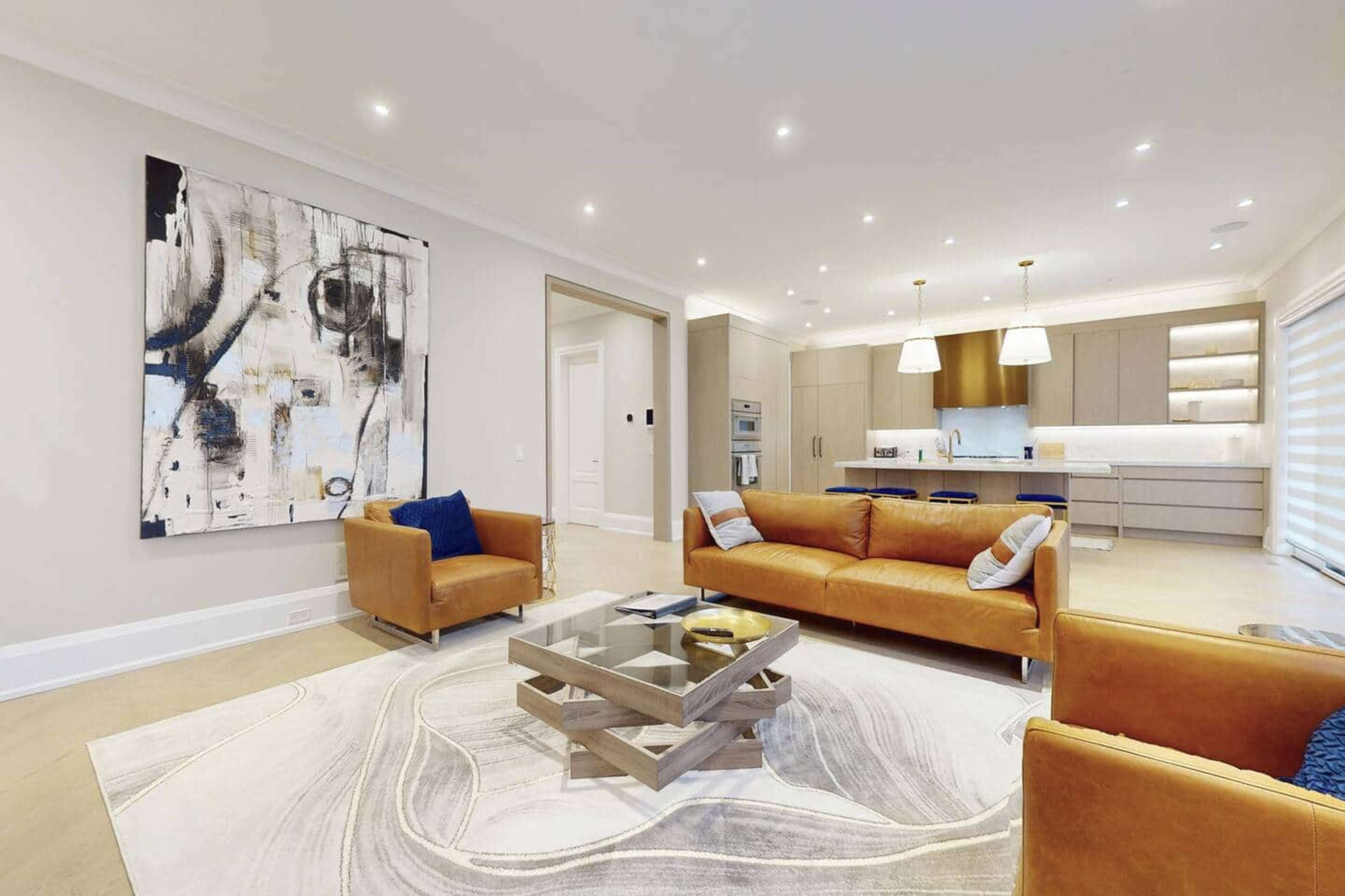 The image shows a modern living room with brown leather sofas, a coffee table, and an abstract artwork on the wall, alongside a kitchen in the background.