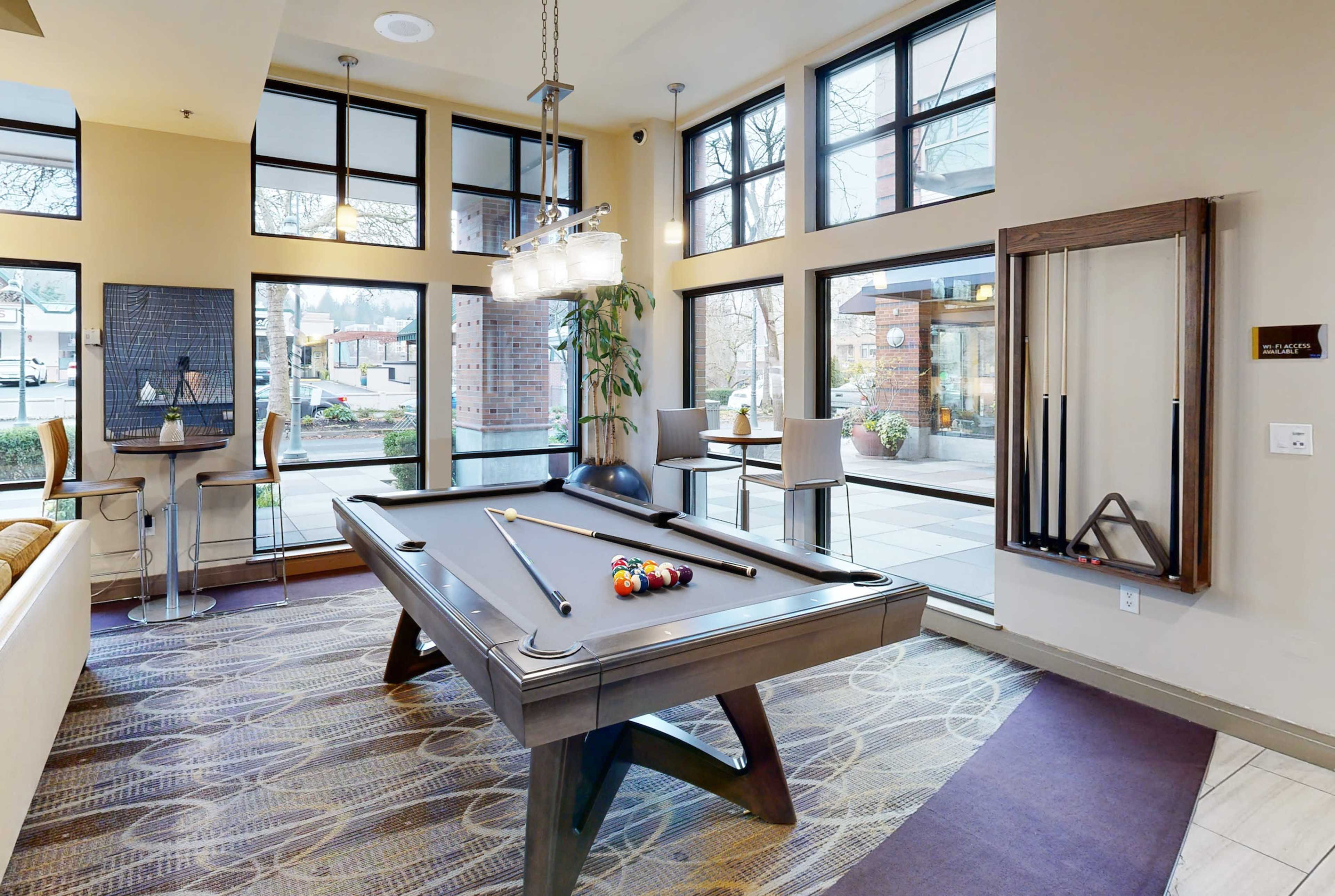 A recreational area with a billiards table in the center, surrounded by large windows and modern seating.