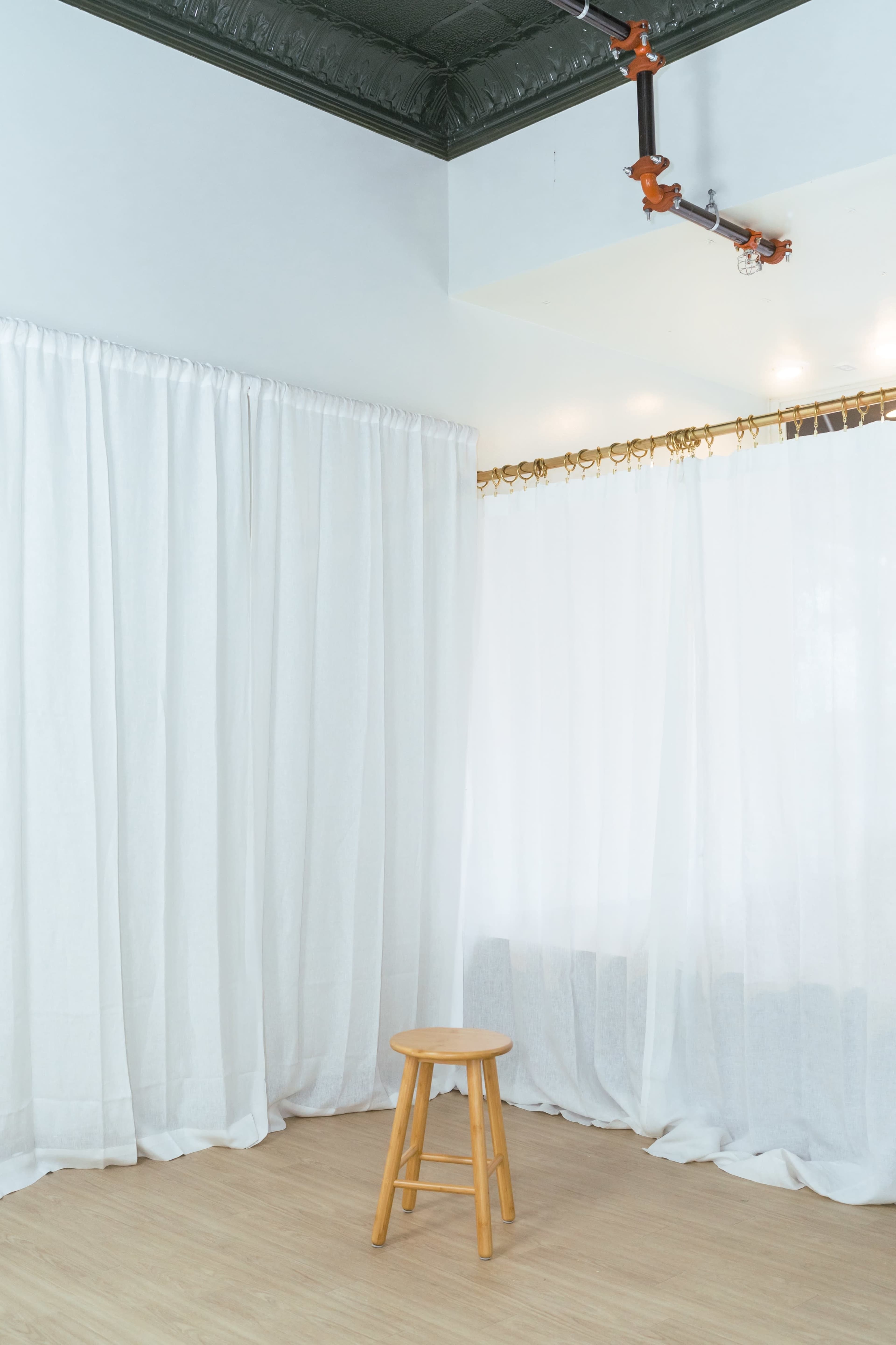 A wooden stool is positioned in a room with white curtains hanging along two walls.