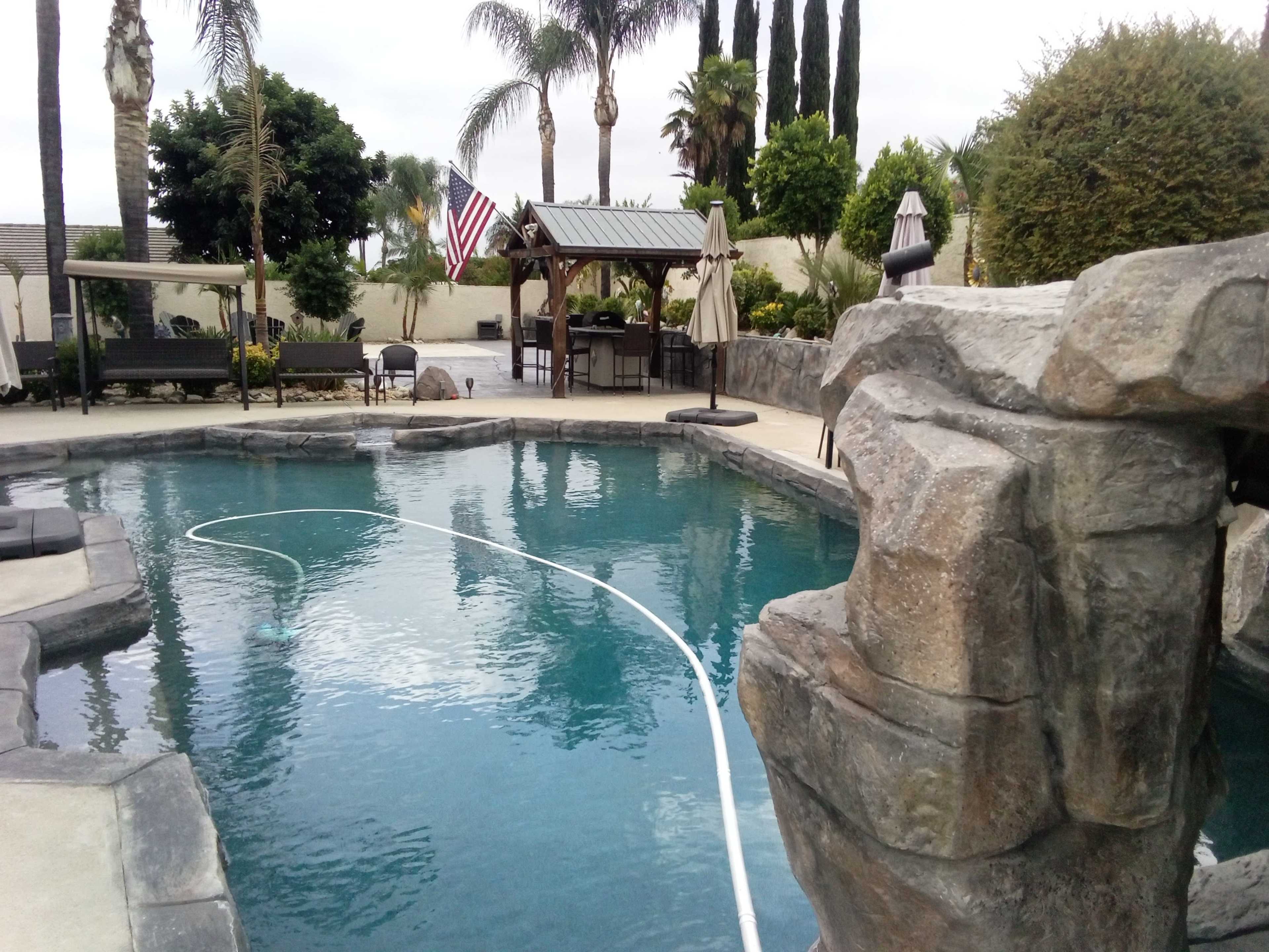 The image shows a backyard swimming pool surrounded by palm trees, lounge chairs, and a gazebo, with an American flag visible in the background.