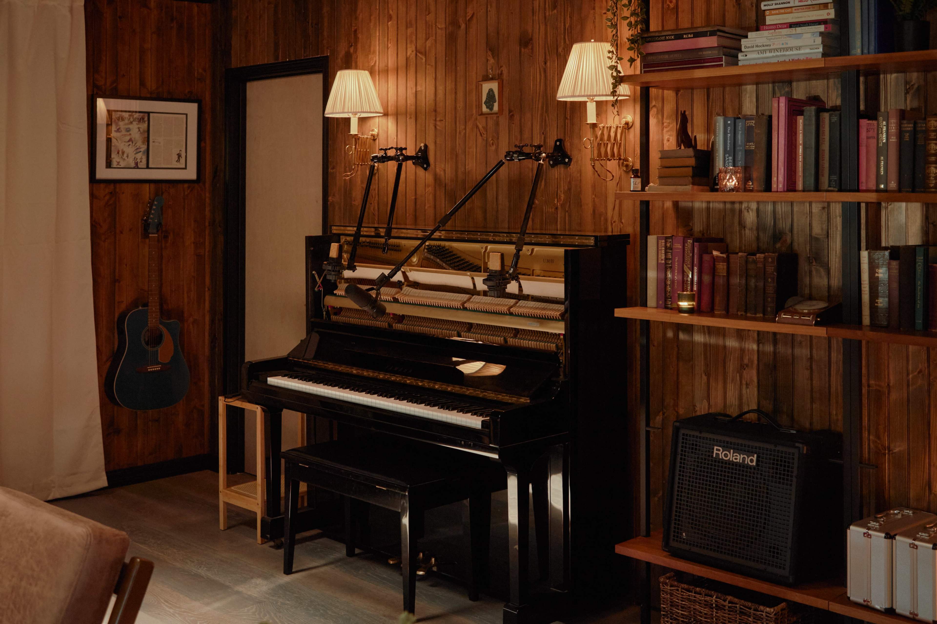 A vintage piano is positioned against a wooden wall, surrounded by bookshelves filled with books, while a guitar leans against the wall.