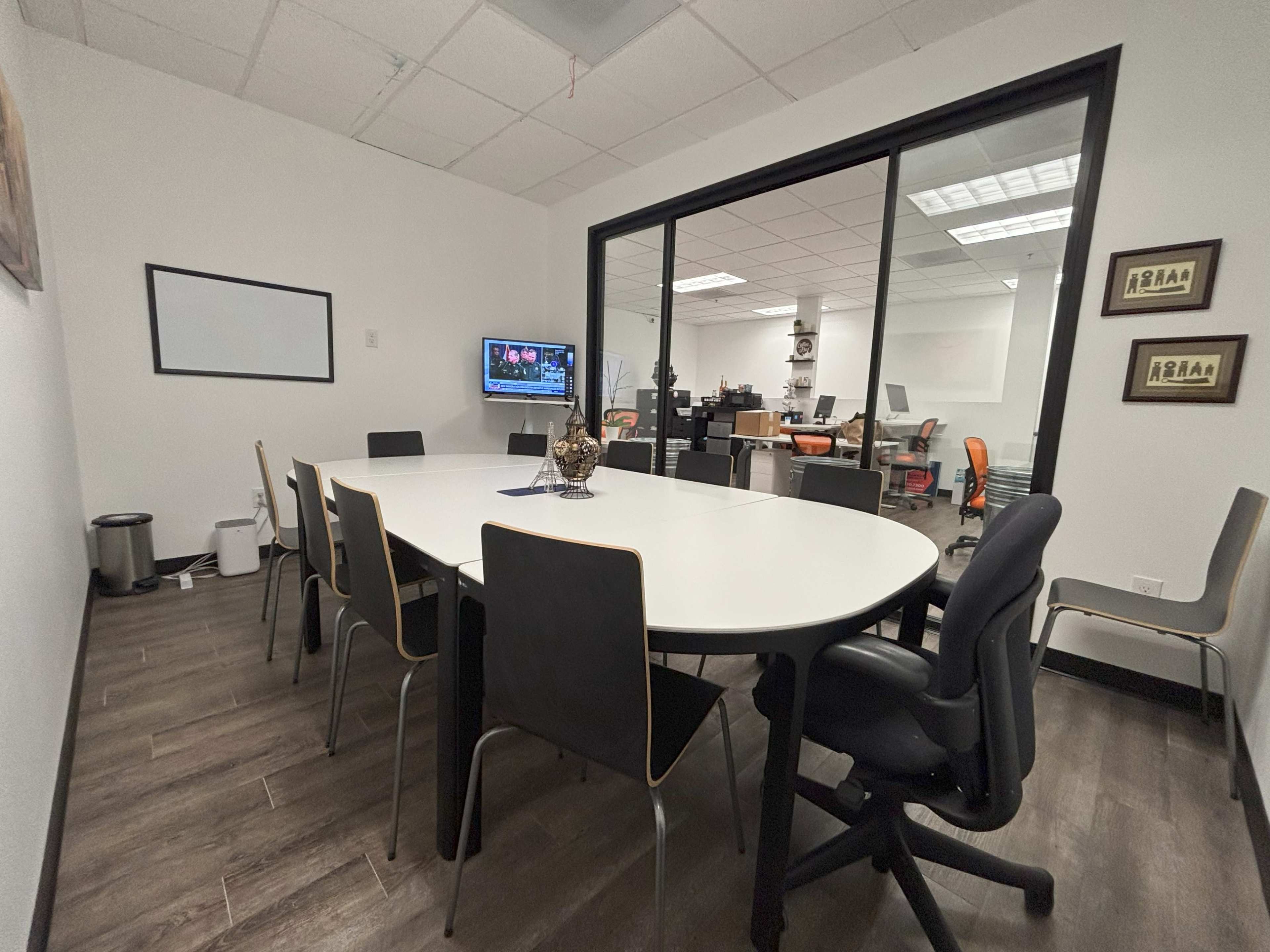 The image shows a conference room with a large round table surrounded by chairs, featuring a television mounted on the wall, and an office space visible through a glass wall.