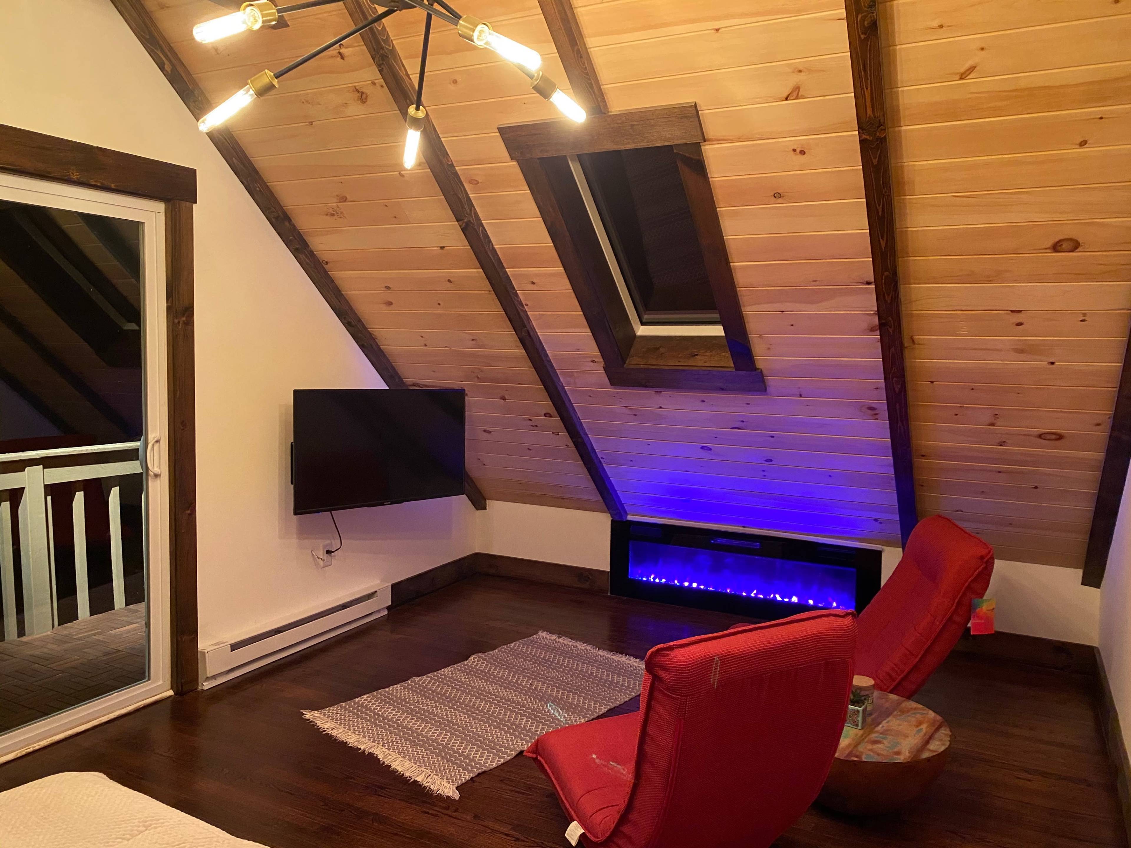 A cozy attic space featuring slanted wooden ceilings, a television mounted on the wall, a low electric fireplace with blue lighting, and two red chairs facing a small table.