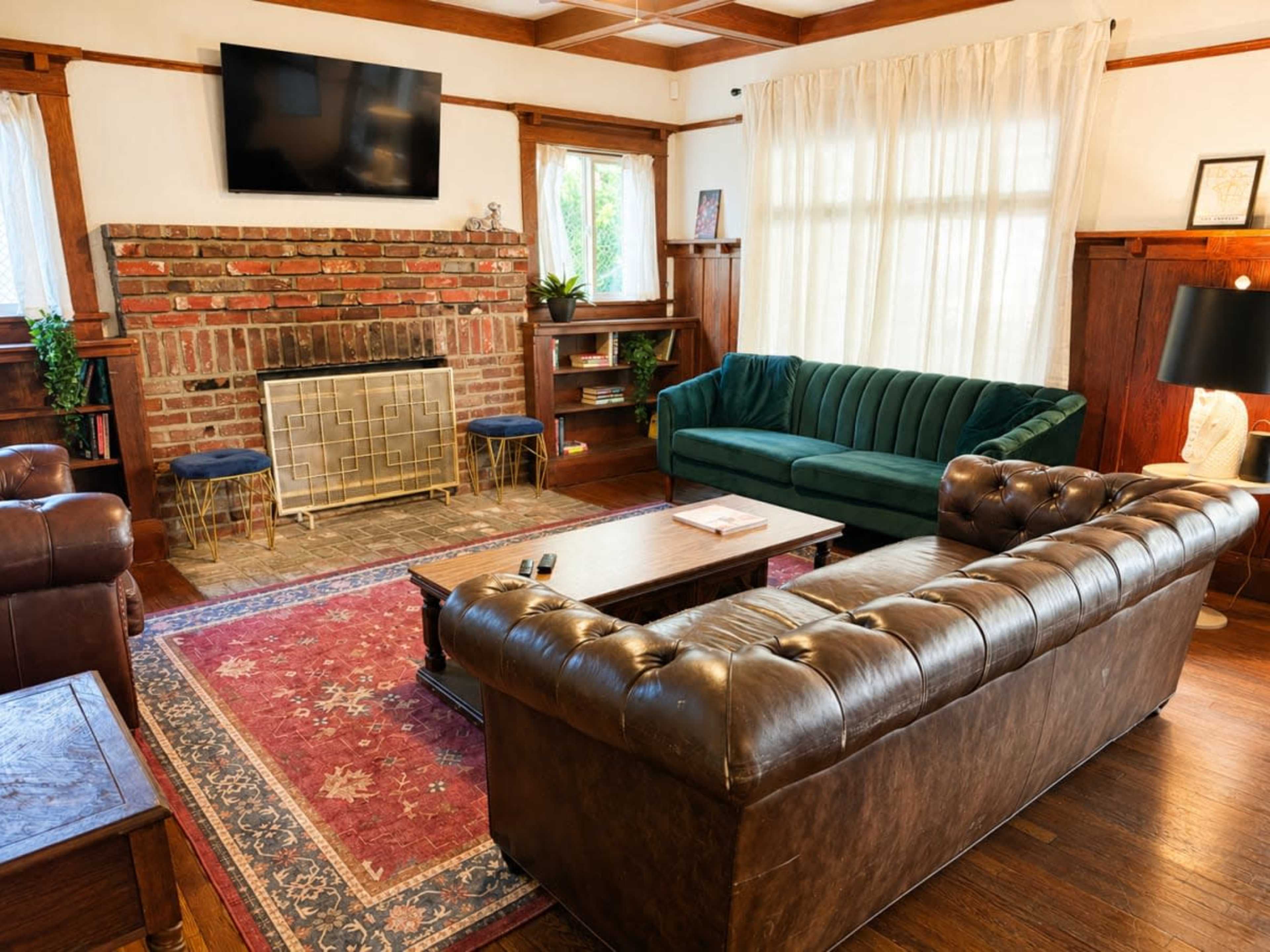 The image shows a cozy living room featuring a brick fireplace, a green velvet sofa, and a brown leather chesterfield couch, with wooden accents and a patterned rug.