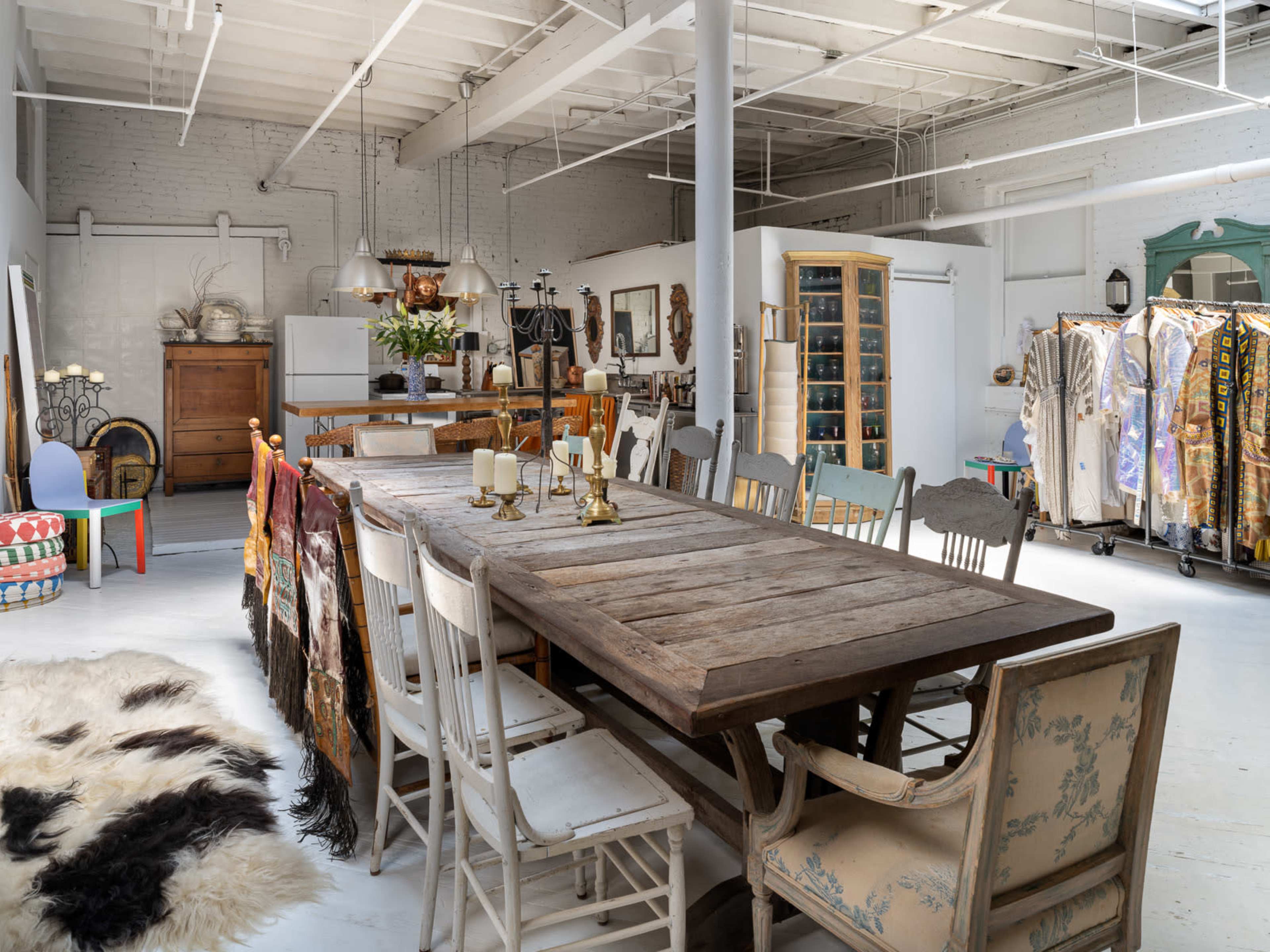 A spacious room with a long wooden dining table surrounded by mismatched chairs, an assortment of clothing on racks, and decorative elements scattered throughout.