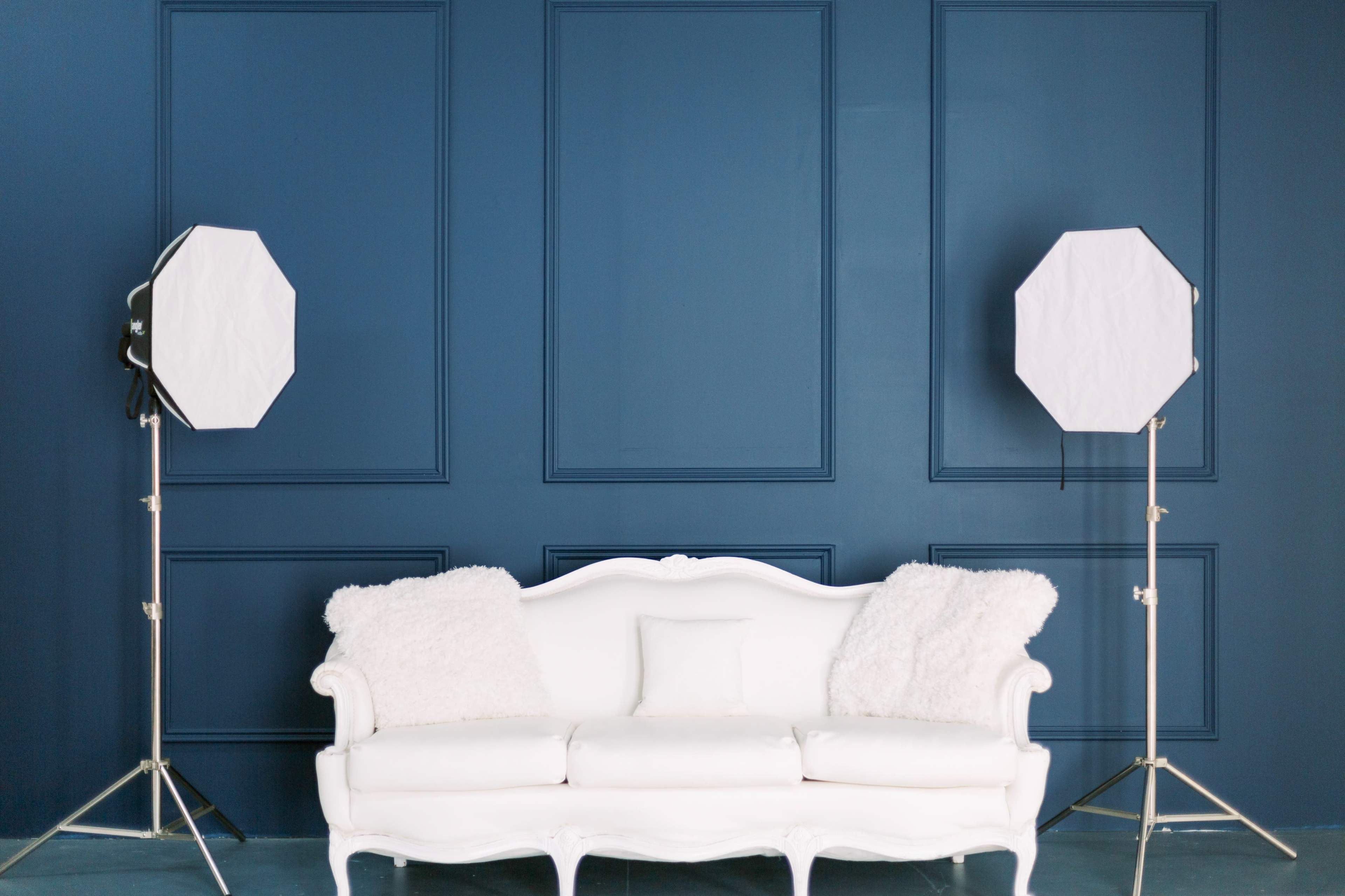 A white couch with two fuzzy pillows is positioned in front of a dark blue wall, flanked by two light stands with softboxes.