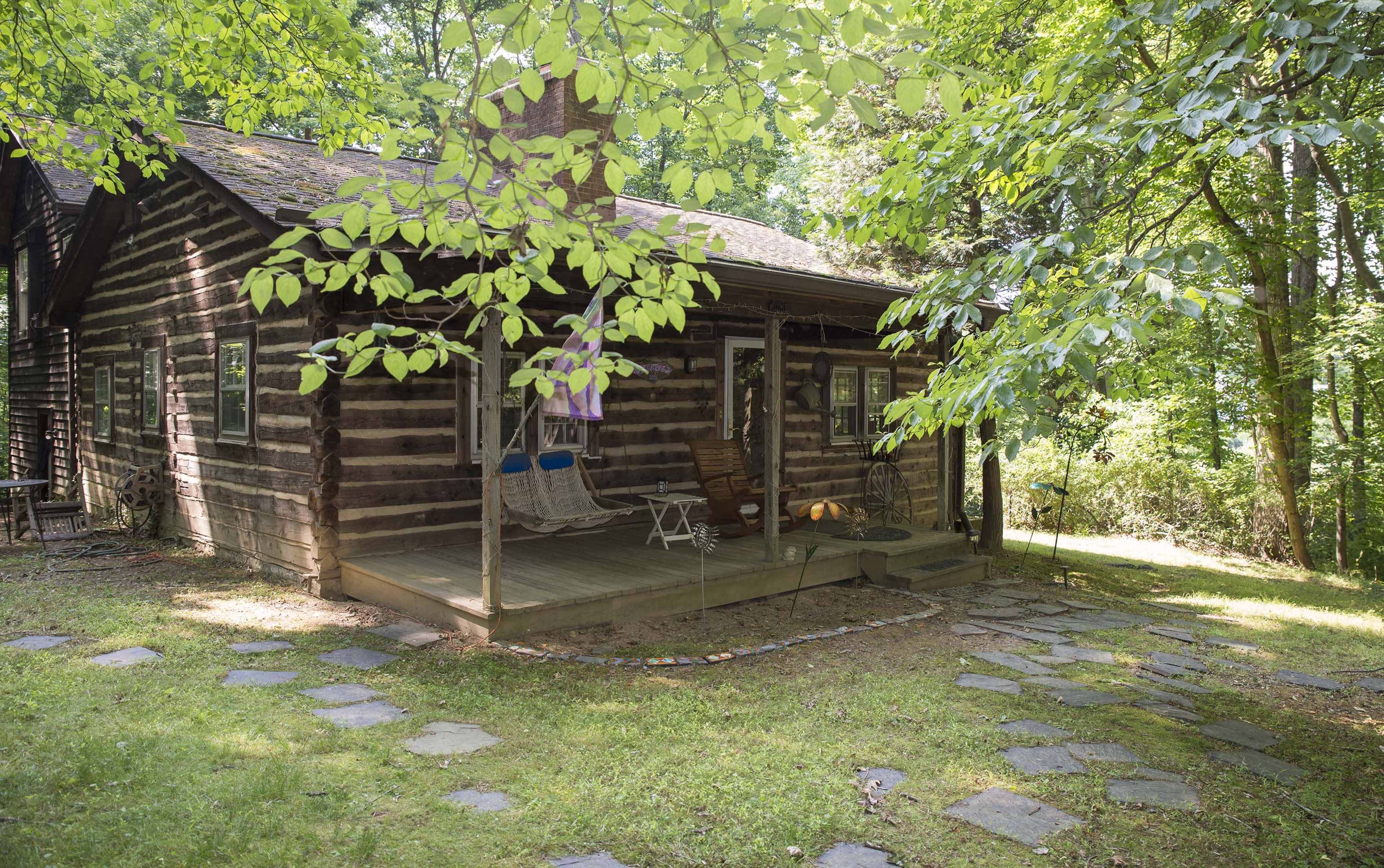 A rustic log cabin is surrounded by lush greenery and features a porch with seating and a stone walkway.