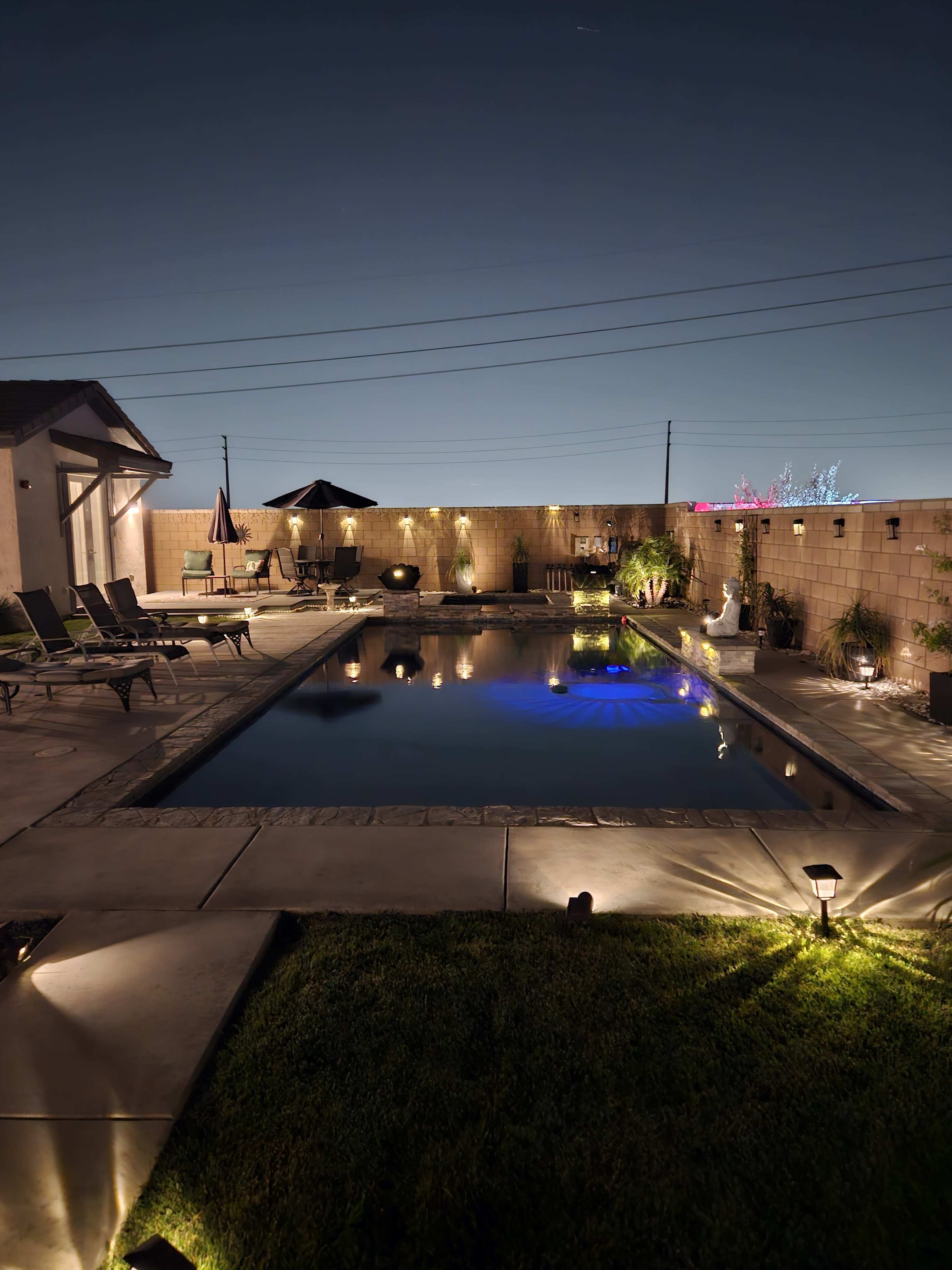 A well-lit backyard features a swimming pool surrounded by loungers and a patio umbrella, set against a clear night sky.