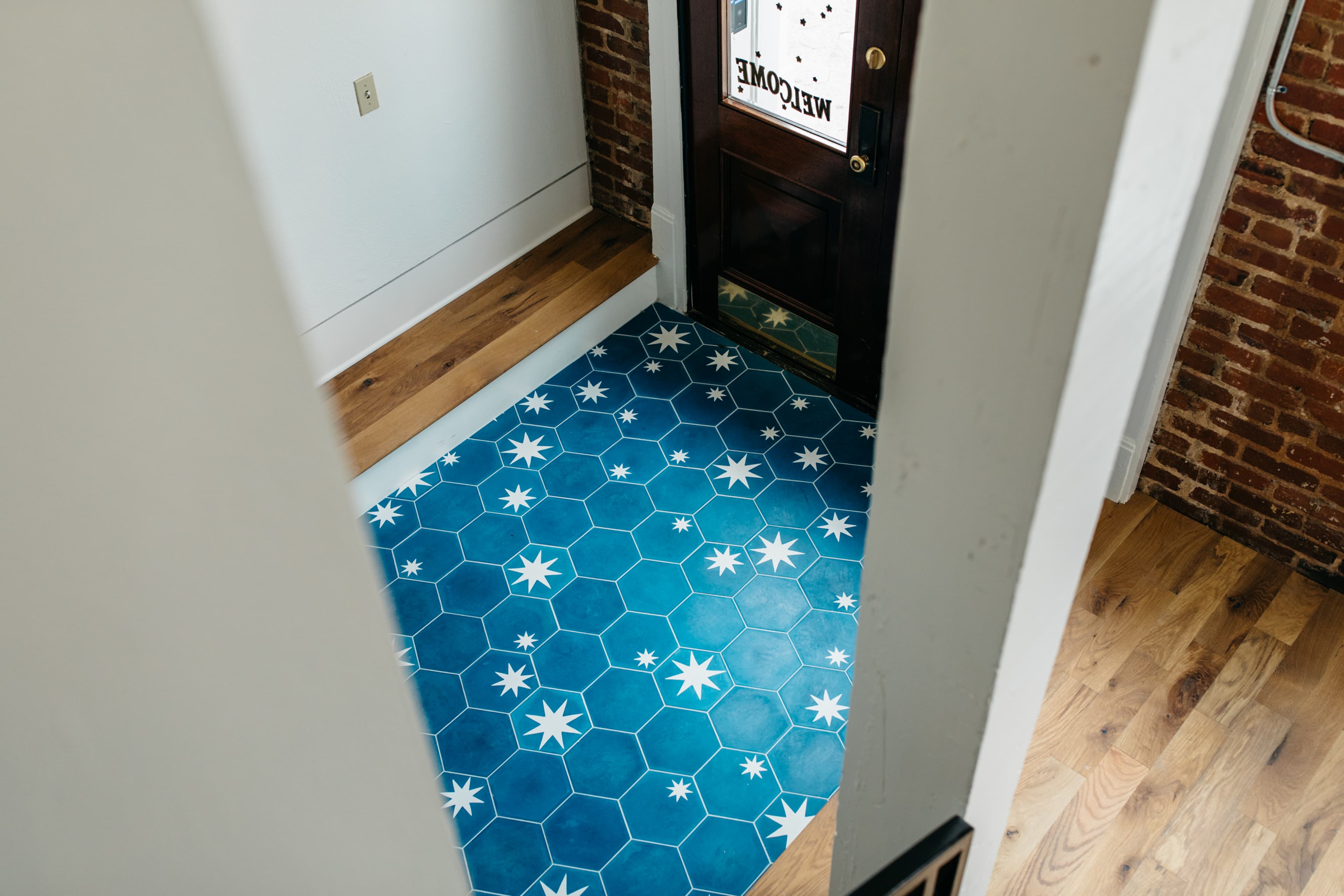 The image shows a hexagonal blue and white tiled floor in an entryway, with a wooden door featuring a "Welcome" sign.