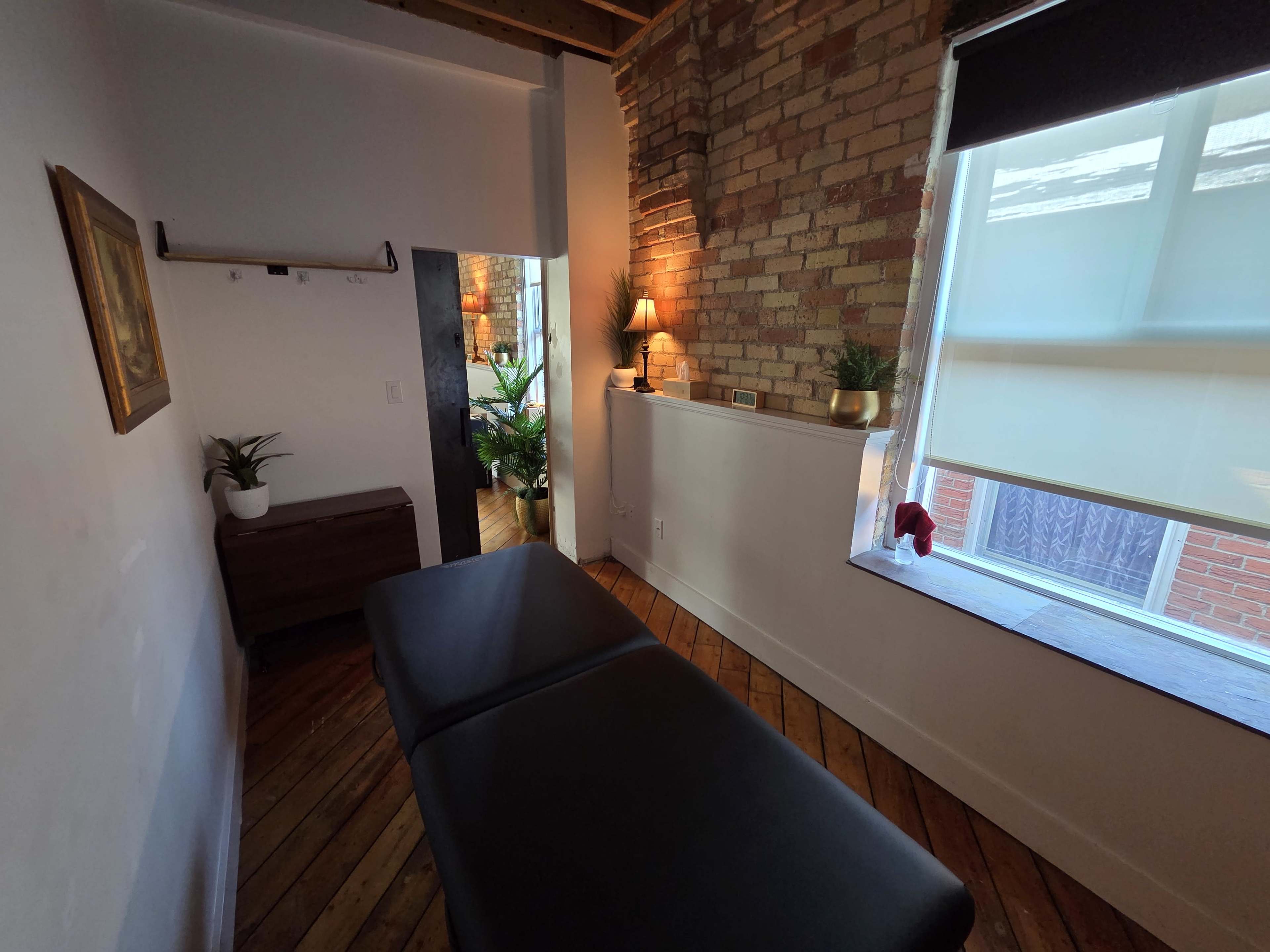 The image shows a massage therapy room with a black treatment table, wooden floors, exposed brick walls, and a window allowing natural light to enter.