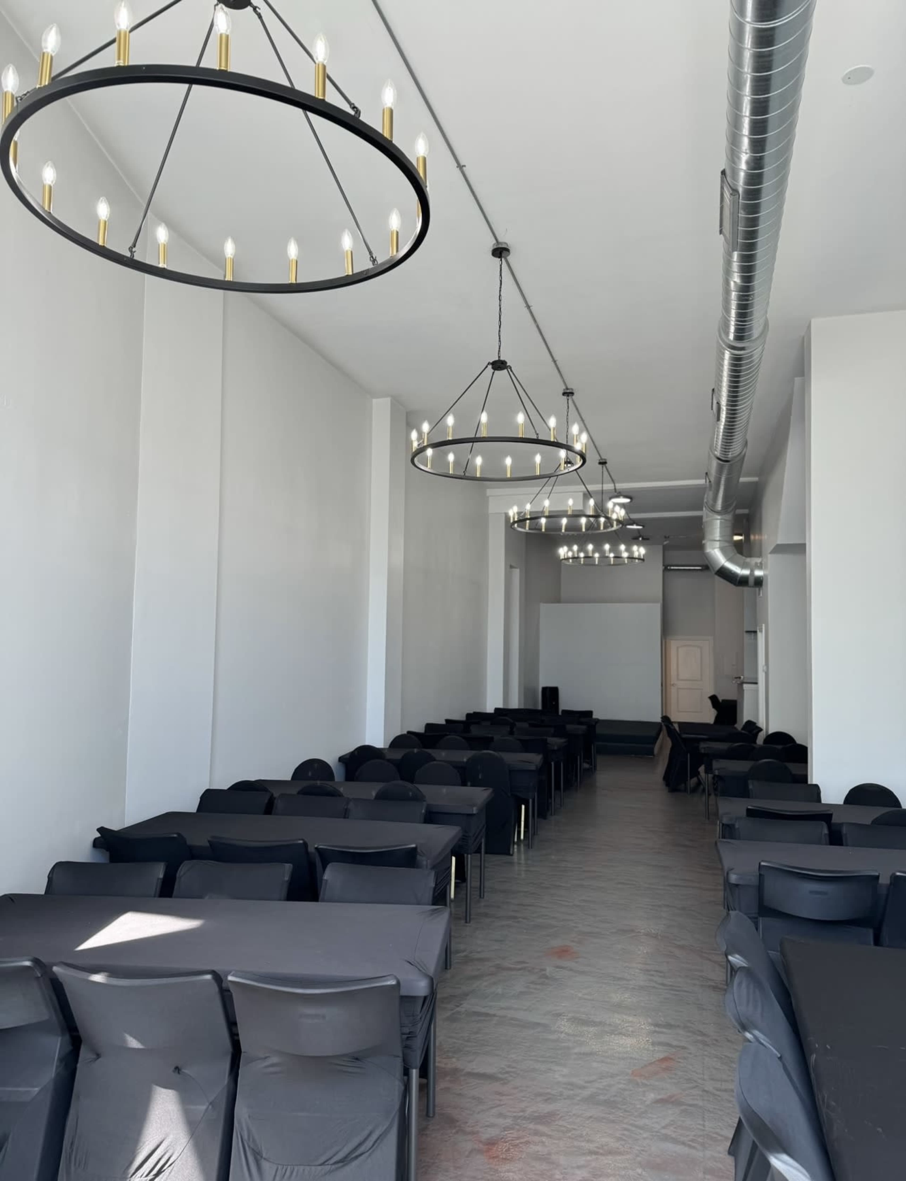 A long, narrow room is set with multiple rows of tables covered in black cloth, featuring circular chandeliers hanging from the ceiling.