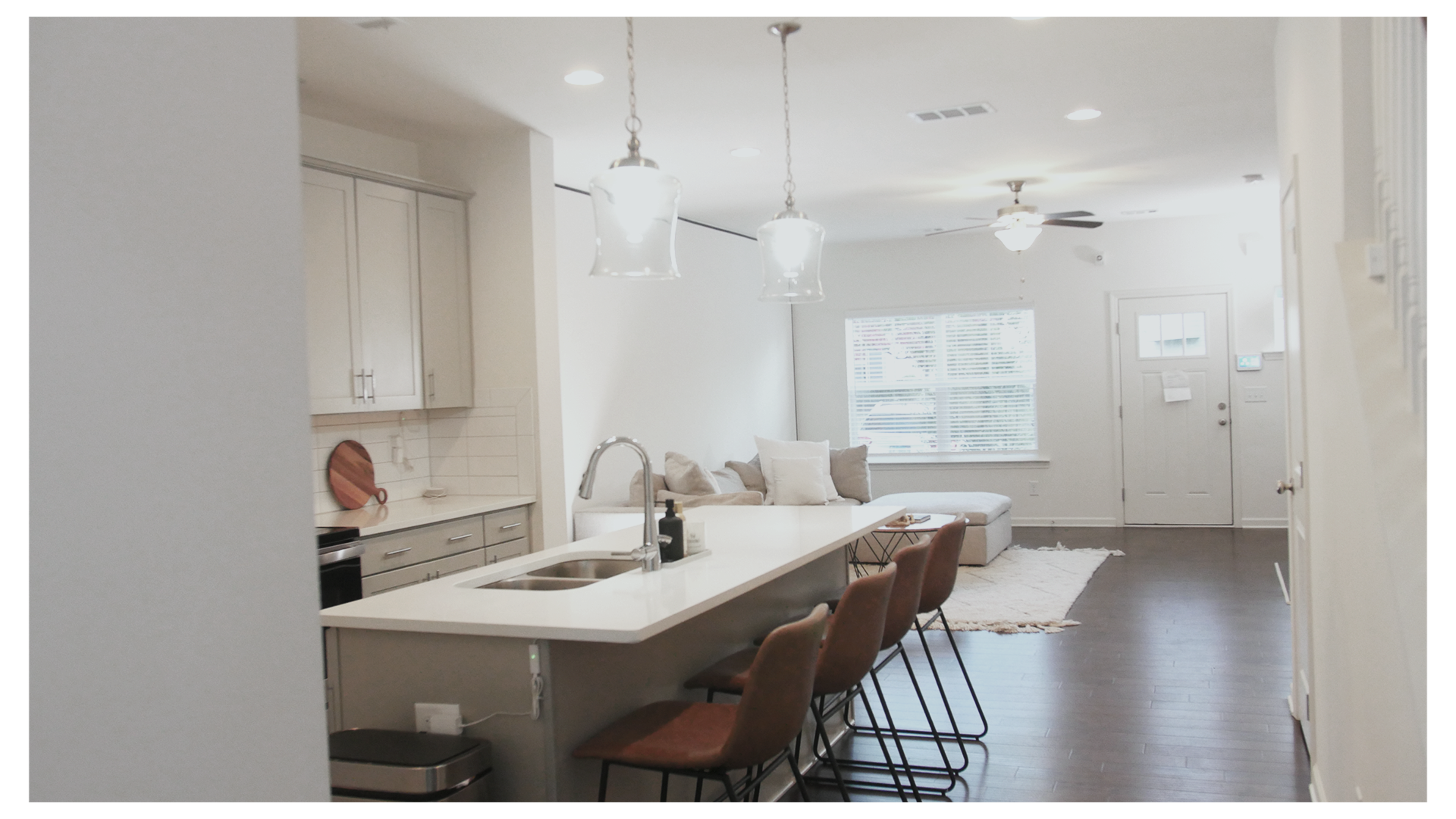 A modern kitchen features gray cabinets, a central island with barstools, and an adjoining living area with a large window and a sectional couch.