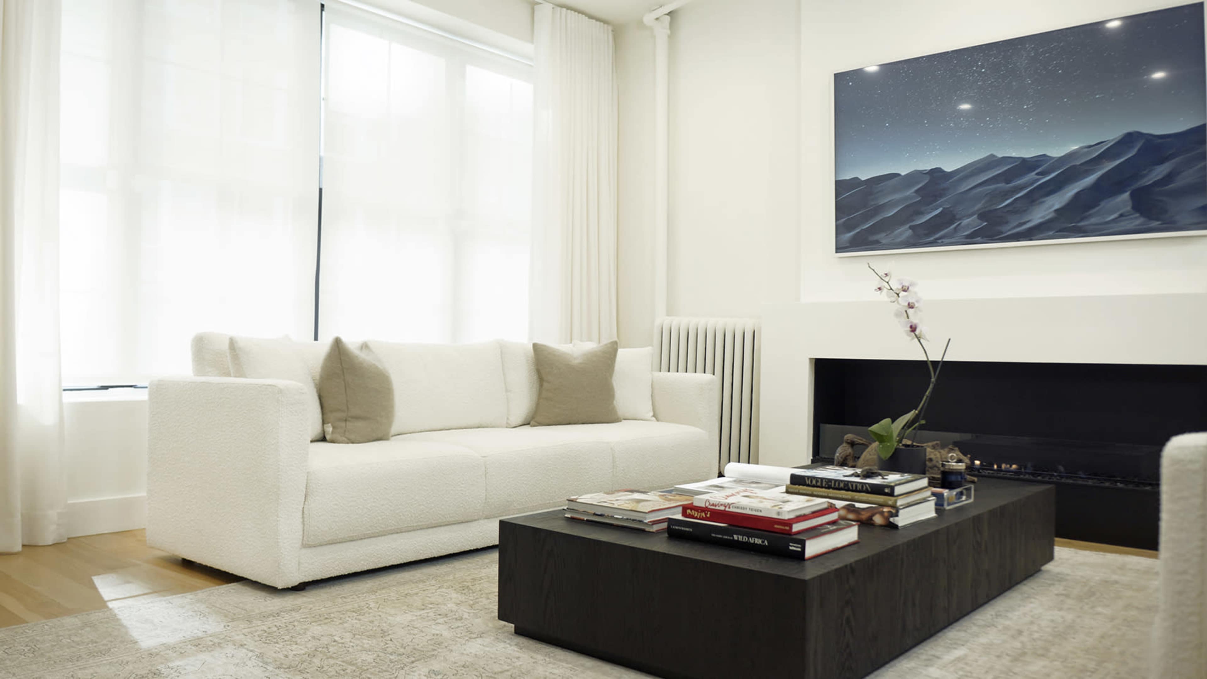 A modern living room features a white sofa, a dark coffee table with books, and an artwork of mountains above a minimalist fireplace.