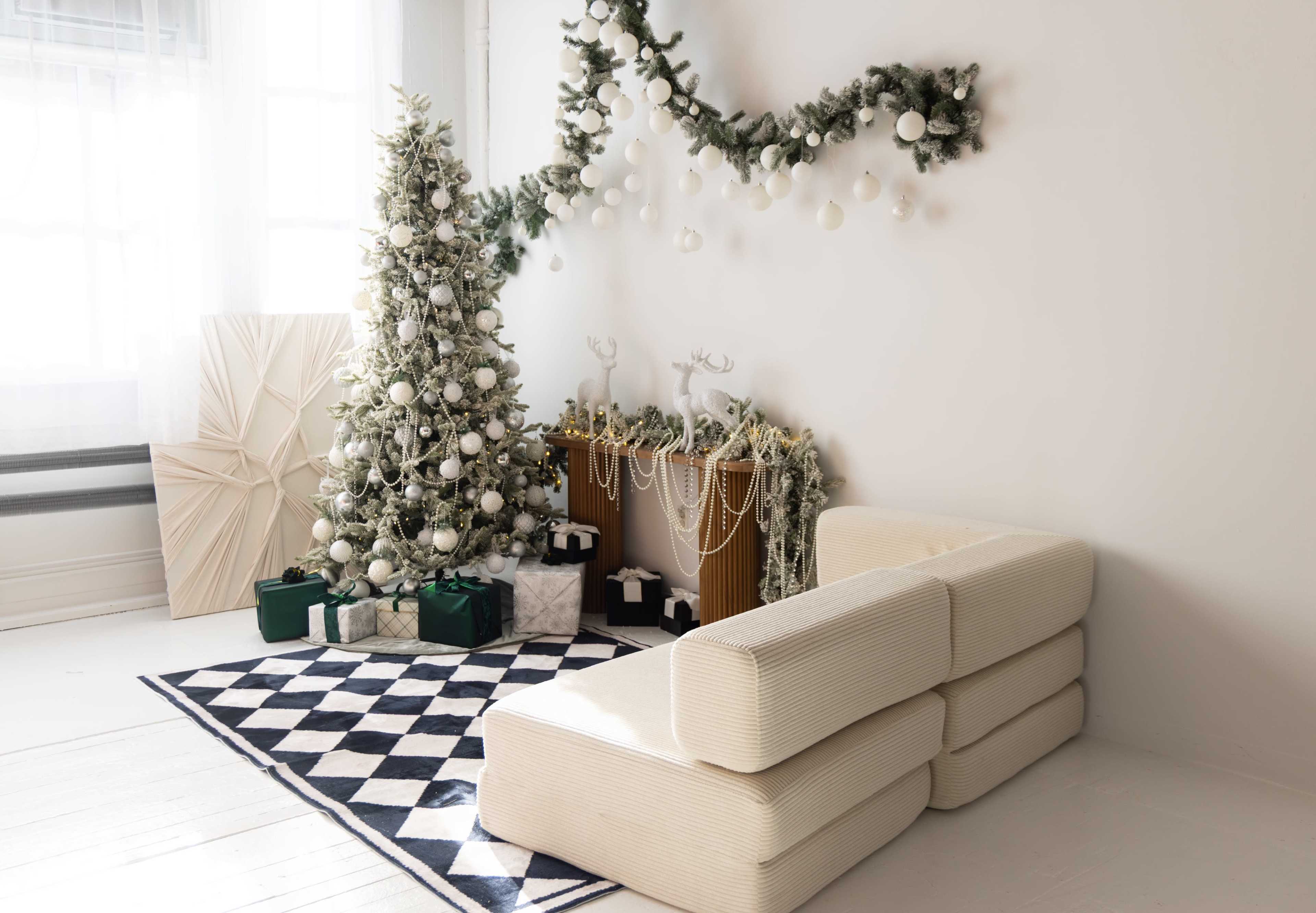 The image features a cozy holiday-themed living space with a decorated Christmas tree, a minimalist white couch, and a festive mantel adorned with garlands and ornaments.