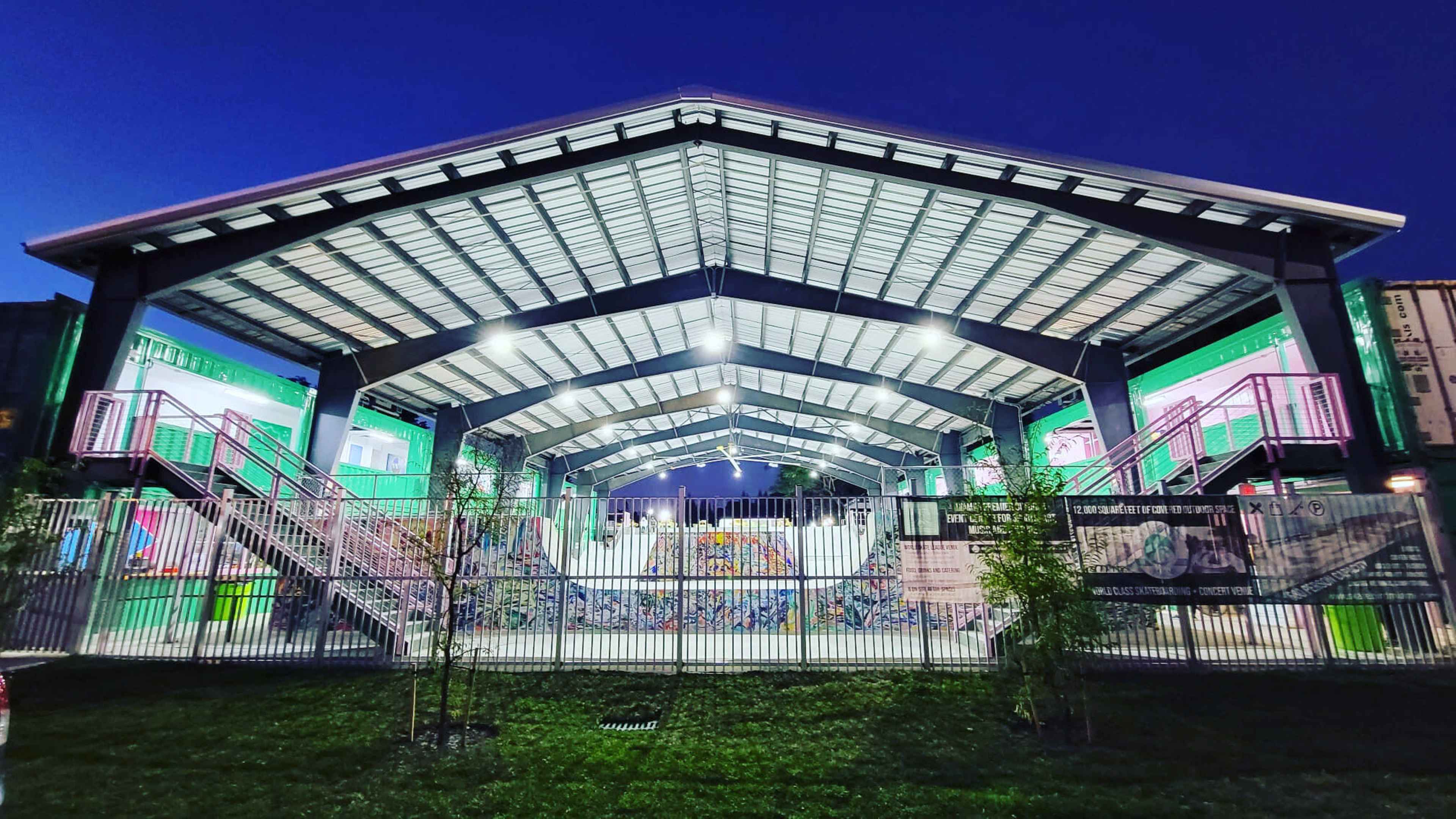 A large, open-air structure with a peaked roof houses a recreational area, illuminated under evening lights, with colorful murals on the walls.