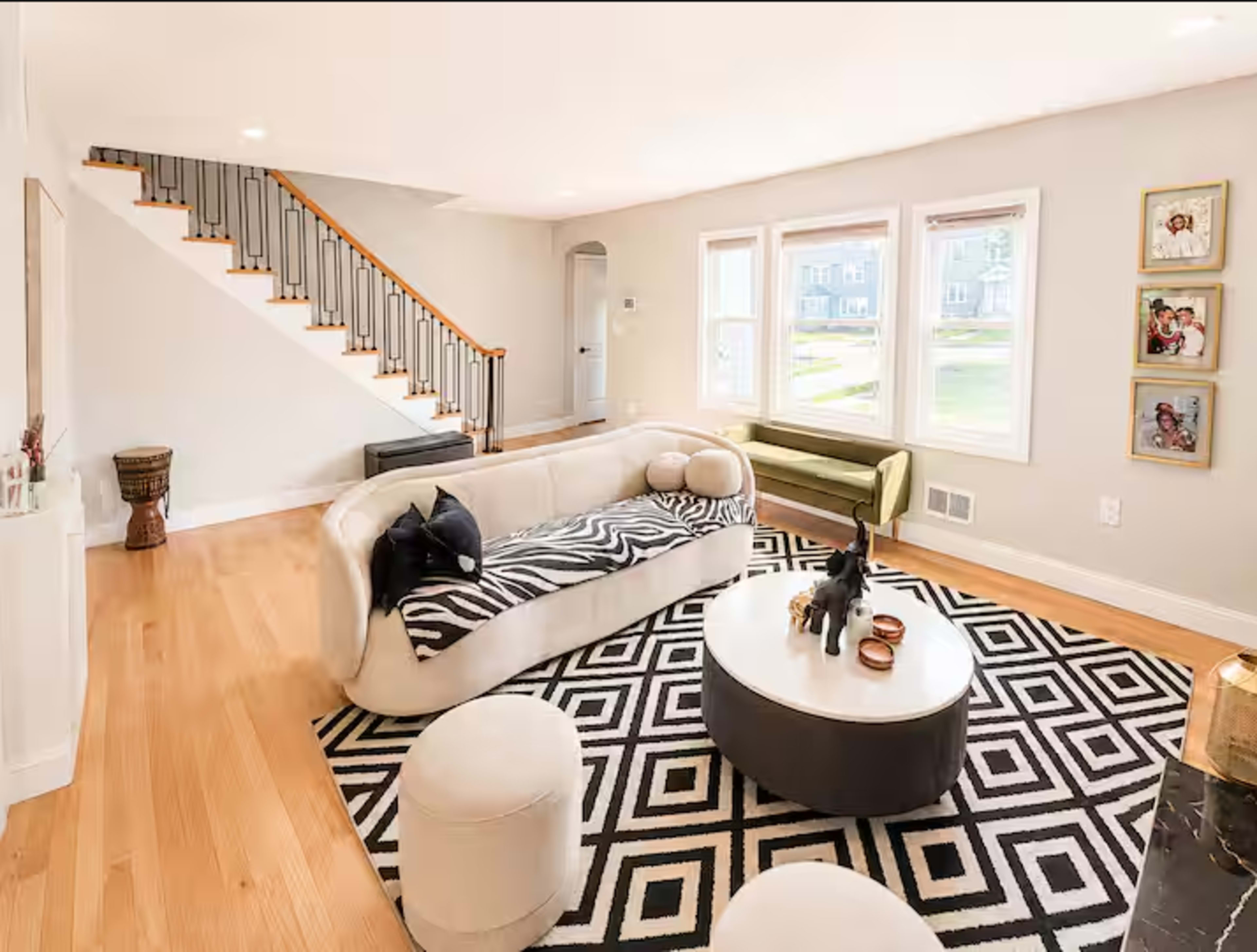 The image shows a modern living room with a light-colored sofa, a patterned rug, and a staircase in the background.