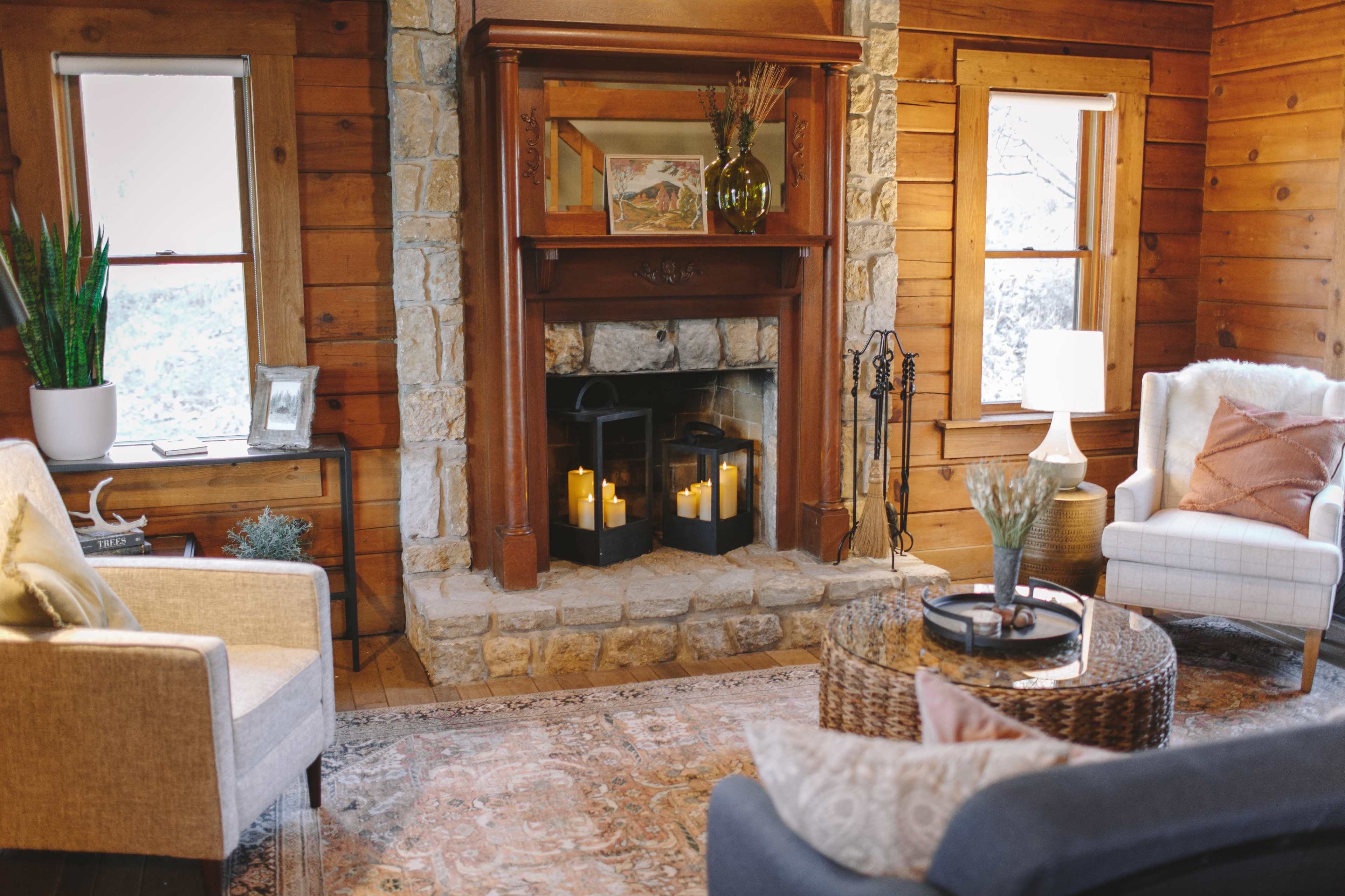 A cozy living room with a stone fireplace, adorned with candles, surrounded by wooden walls and comfortable seating.