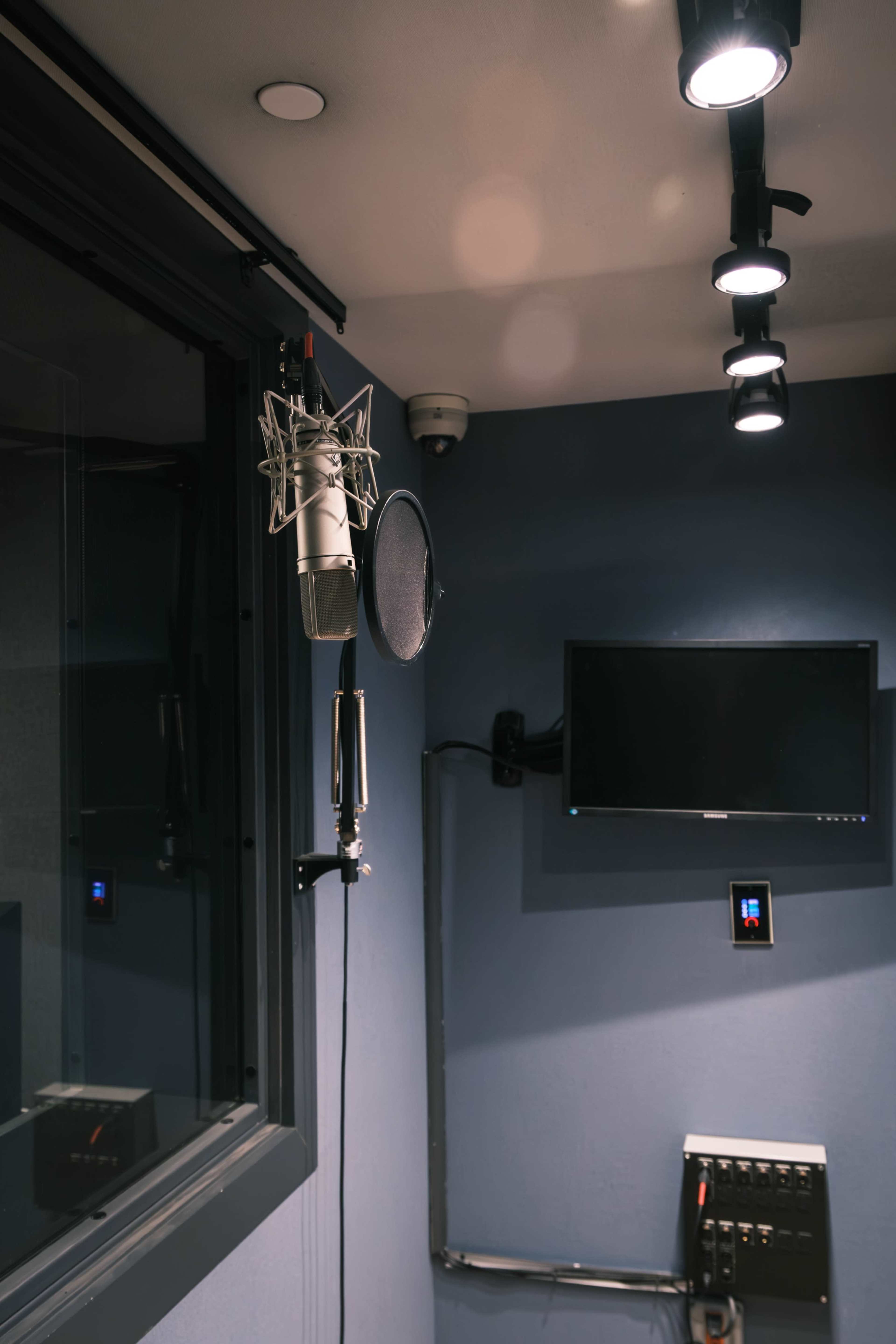 A microphone and pop filter are set up in a soundproof recording booth, with a television mounted on the wall.