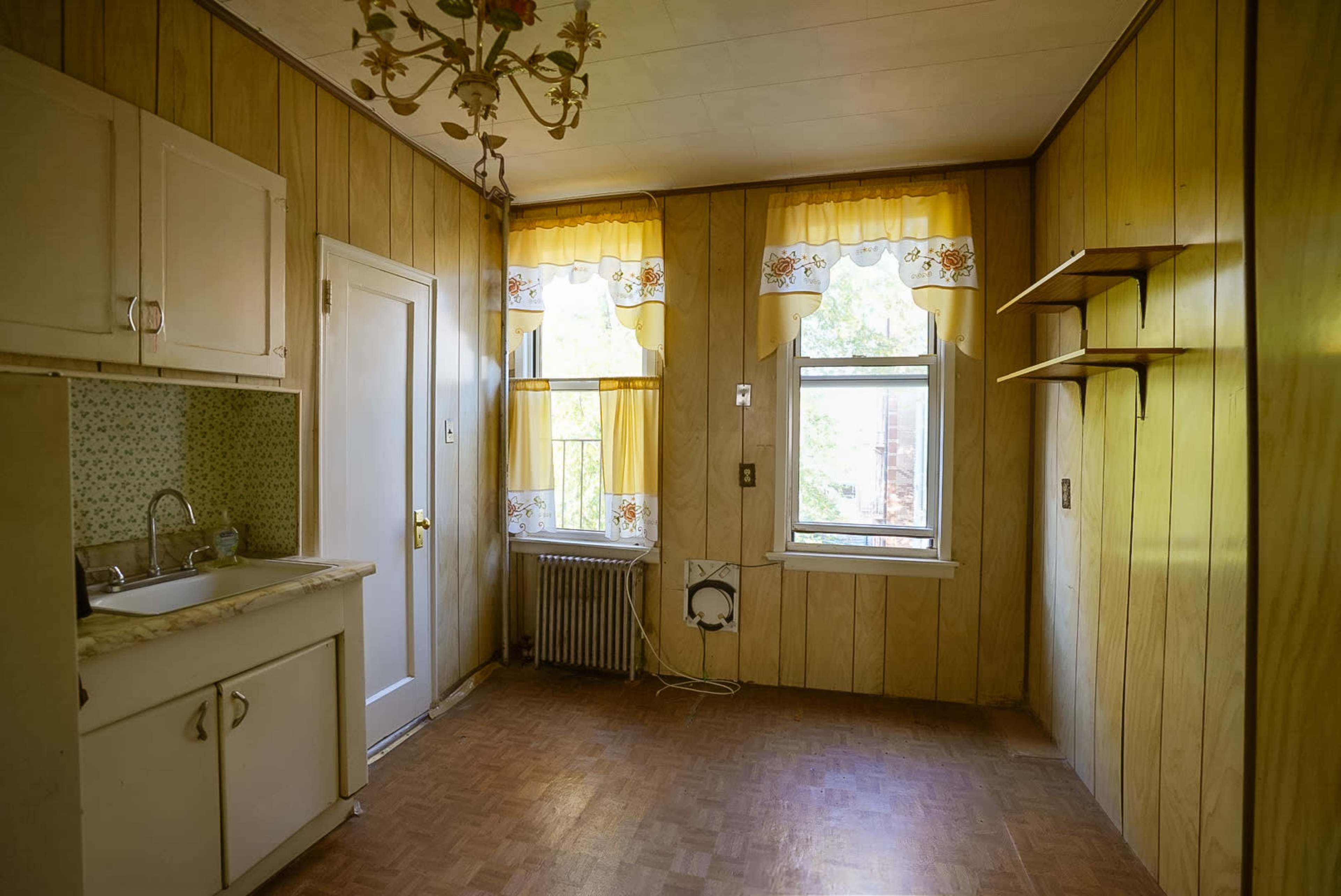 The image shows a small, light-filled kitchen with wooden paneling, a sink on the left, and yellow curtains at the window.