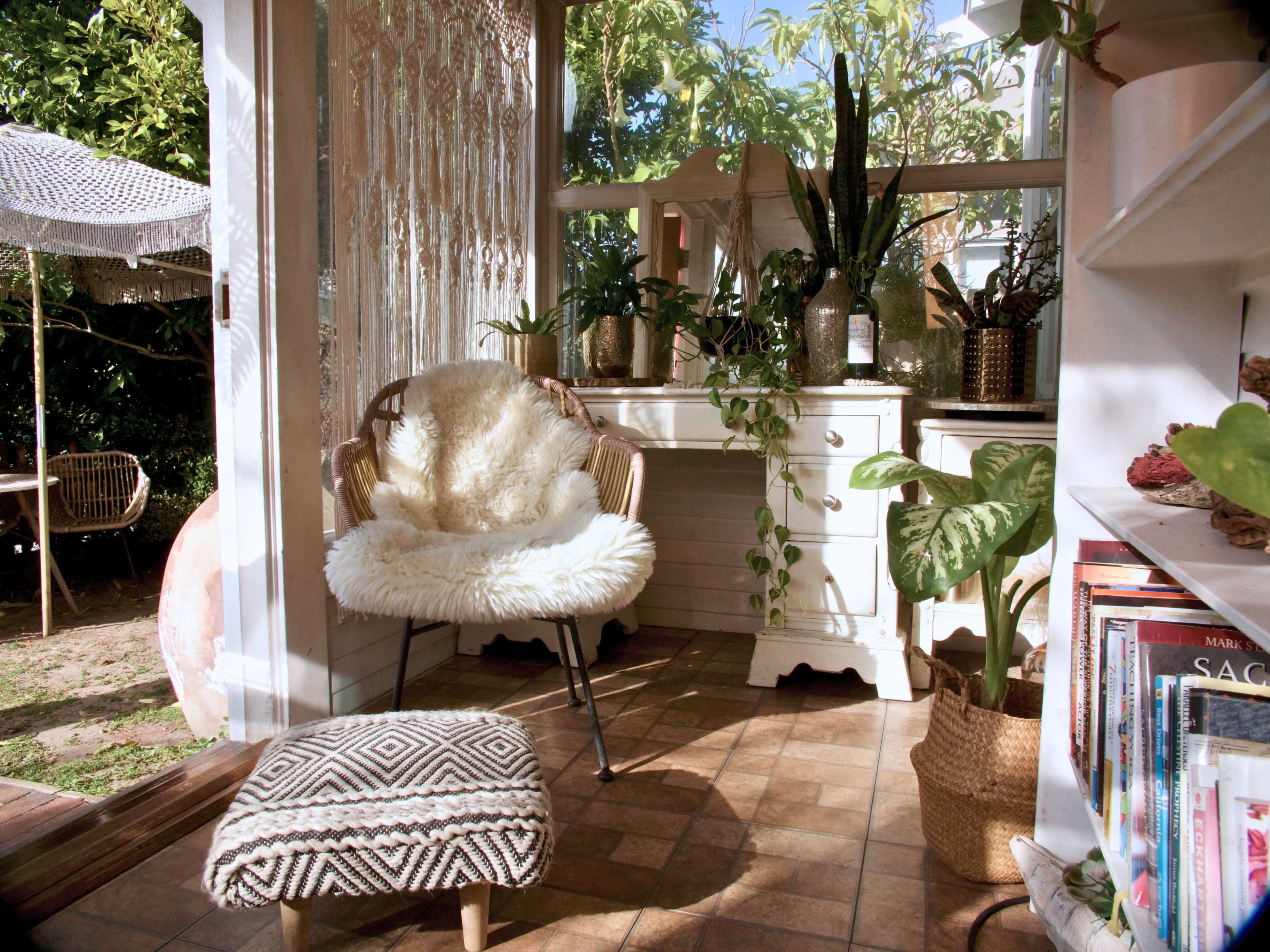 The image shows a cozy indoor corner with a soft chair and a small pouf, surrounded by plants and a white chest of drawers.