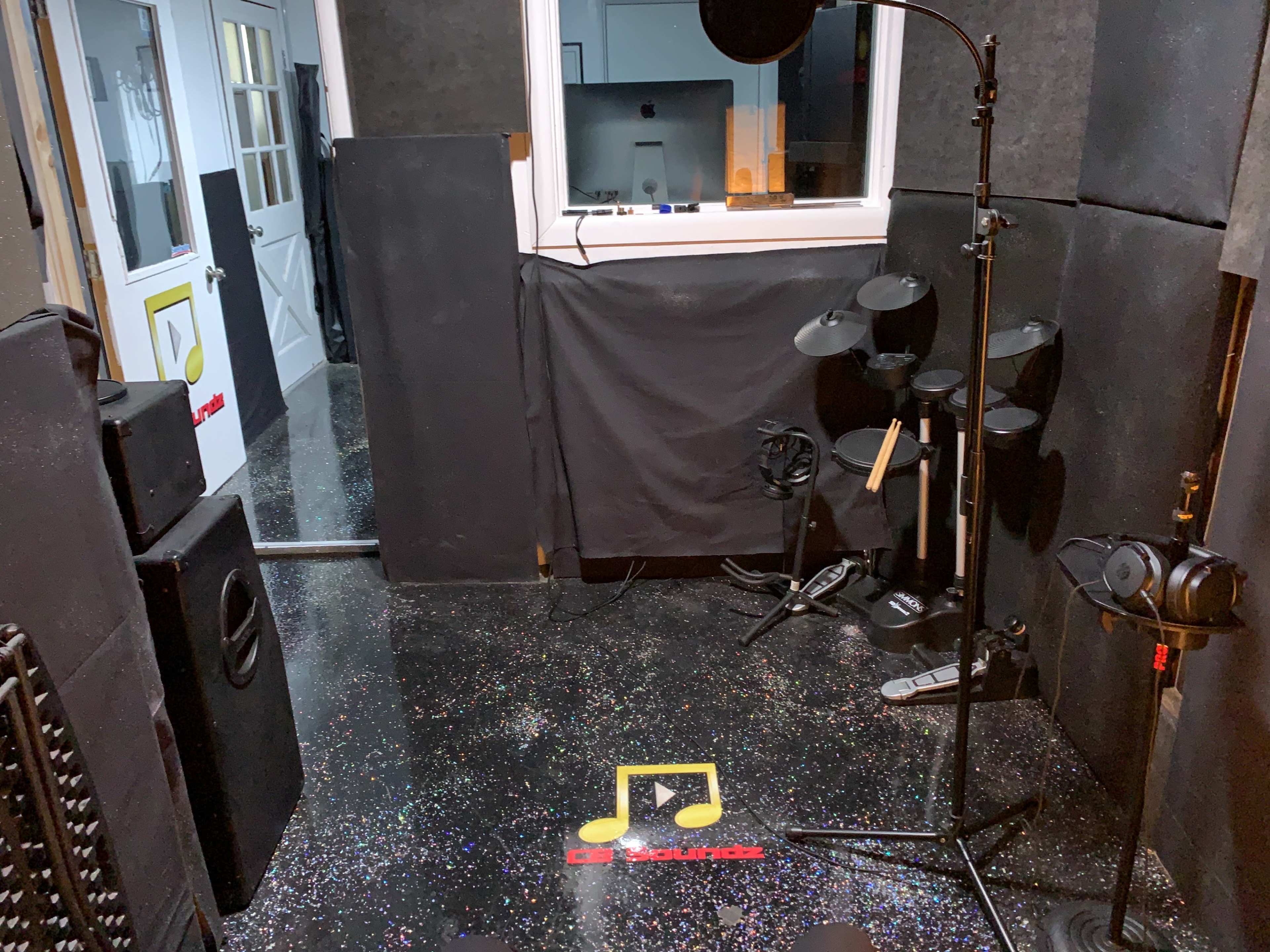 The image shows a soundproof recording studio with a microphone stand, electronic drums, and sound equipment against dark padded walls.