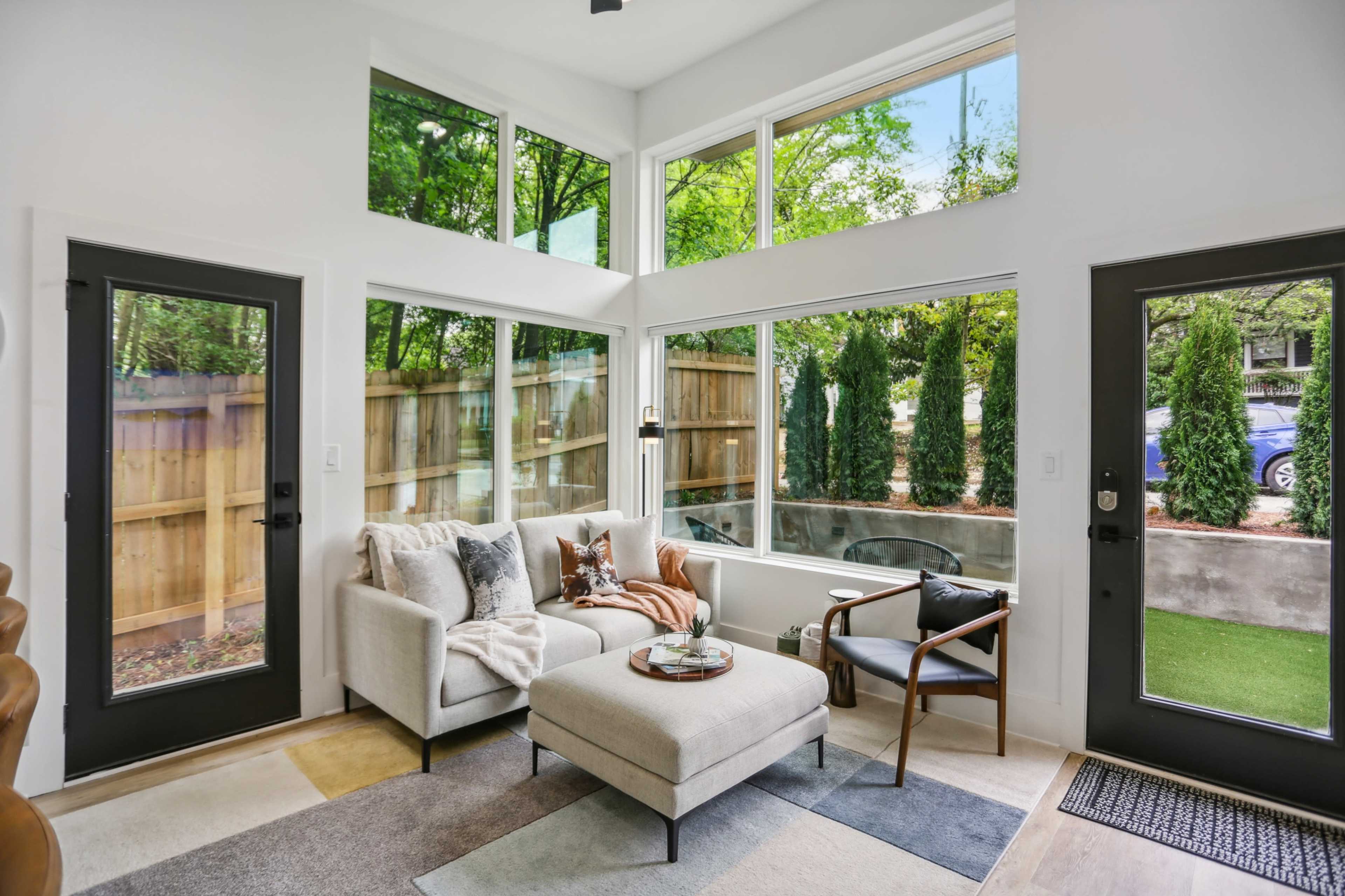 A modern living room features large windows, a light-colored sofa, and a small coffee table, with views of a green outdoor area.
