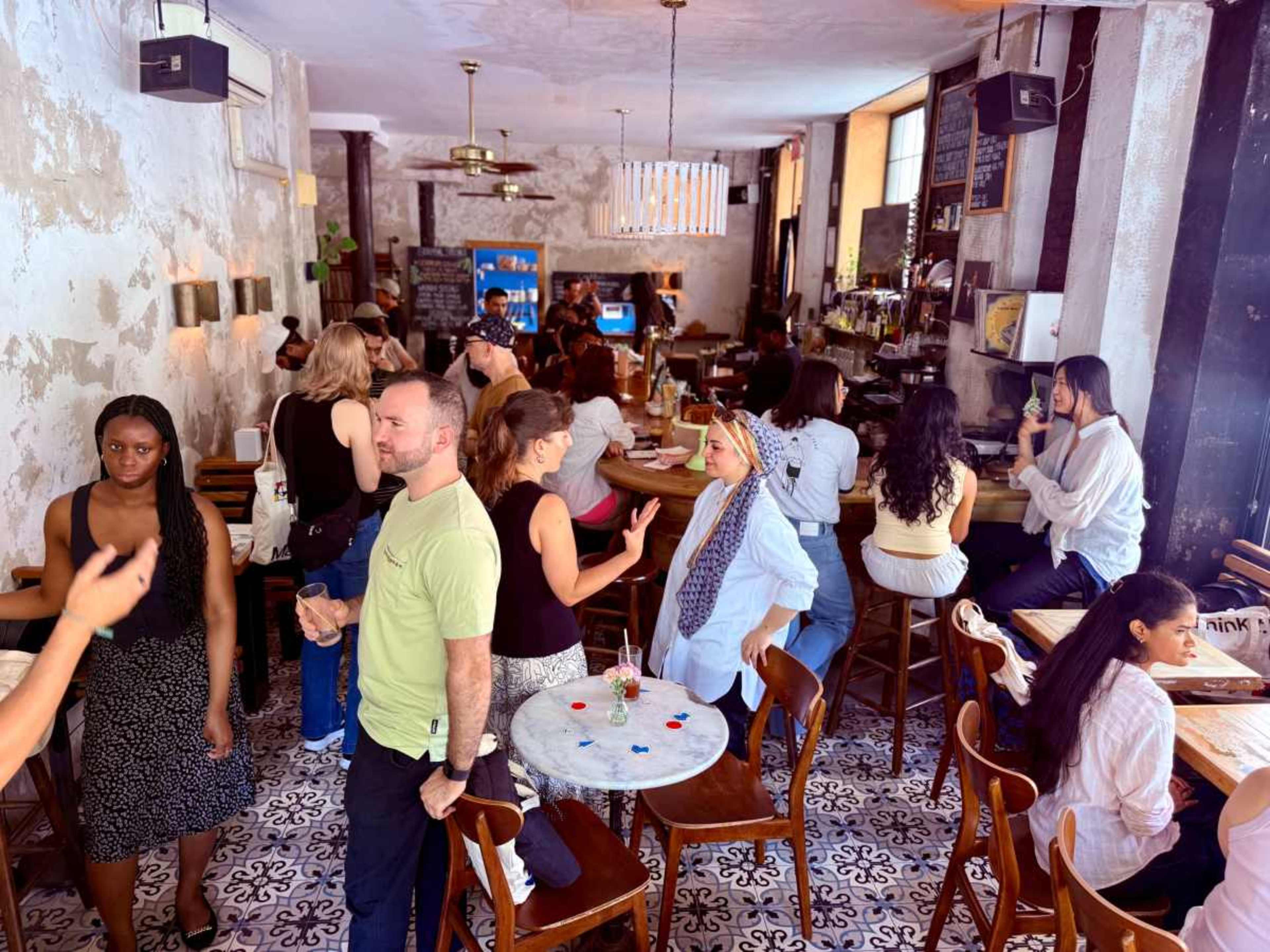 The image shows a bustling café filled with people seated at tables and standing around a bar, with a mix of styles and activities among the patrons.