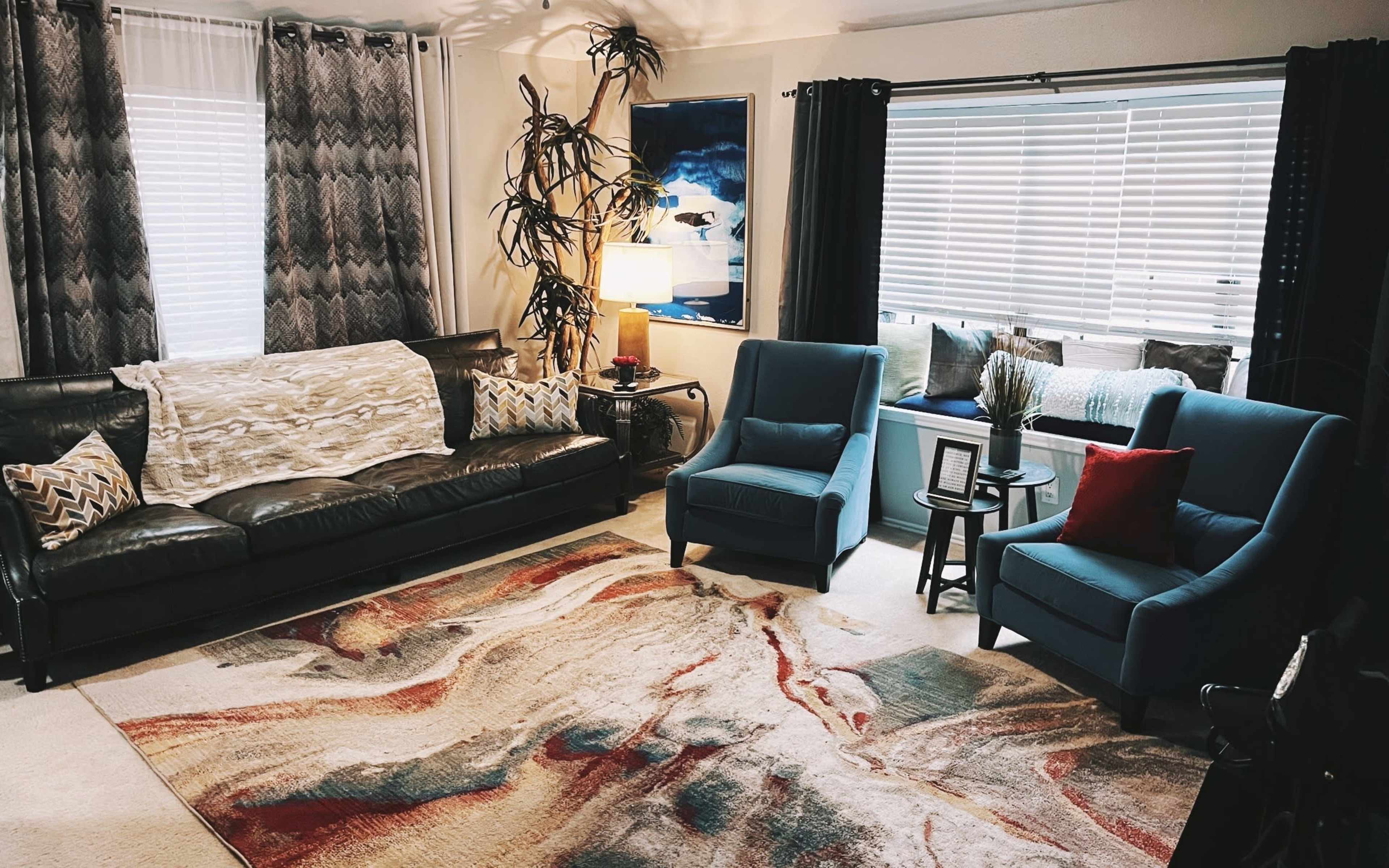 A living room with a black leather sofa, two blue chairs, a large area rug, and a window seat with cushions.