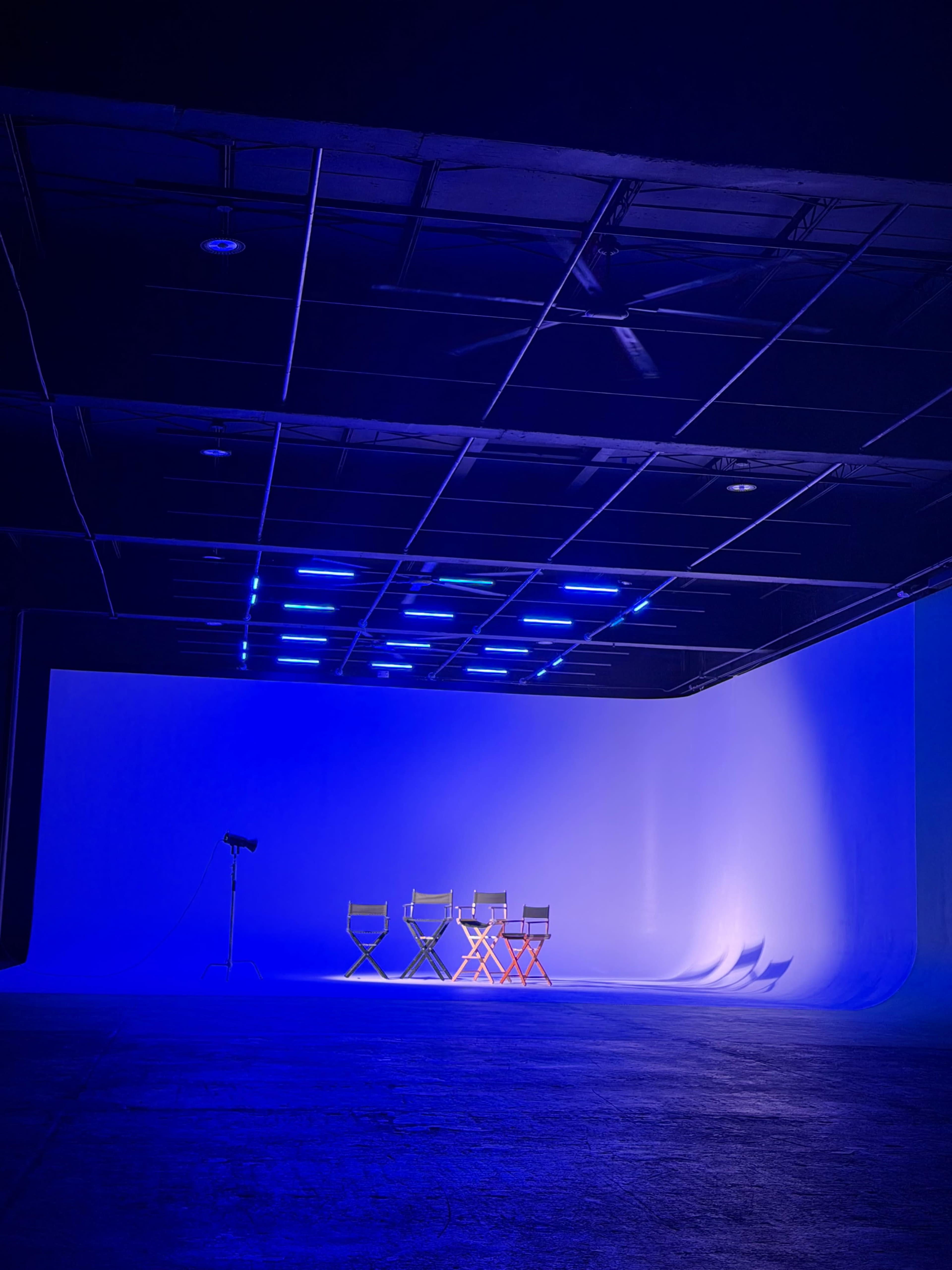 The image shows a large, empty studio space with a blue backdrop and three director's chairs, two gray and one orange, illuminated by blue lights above.