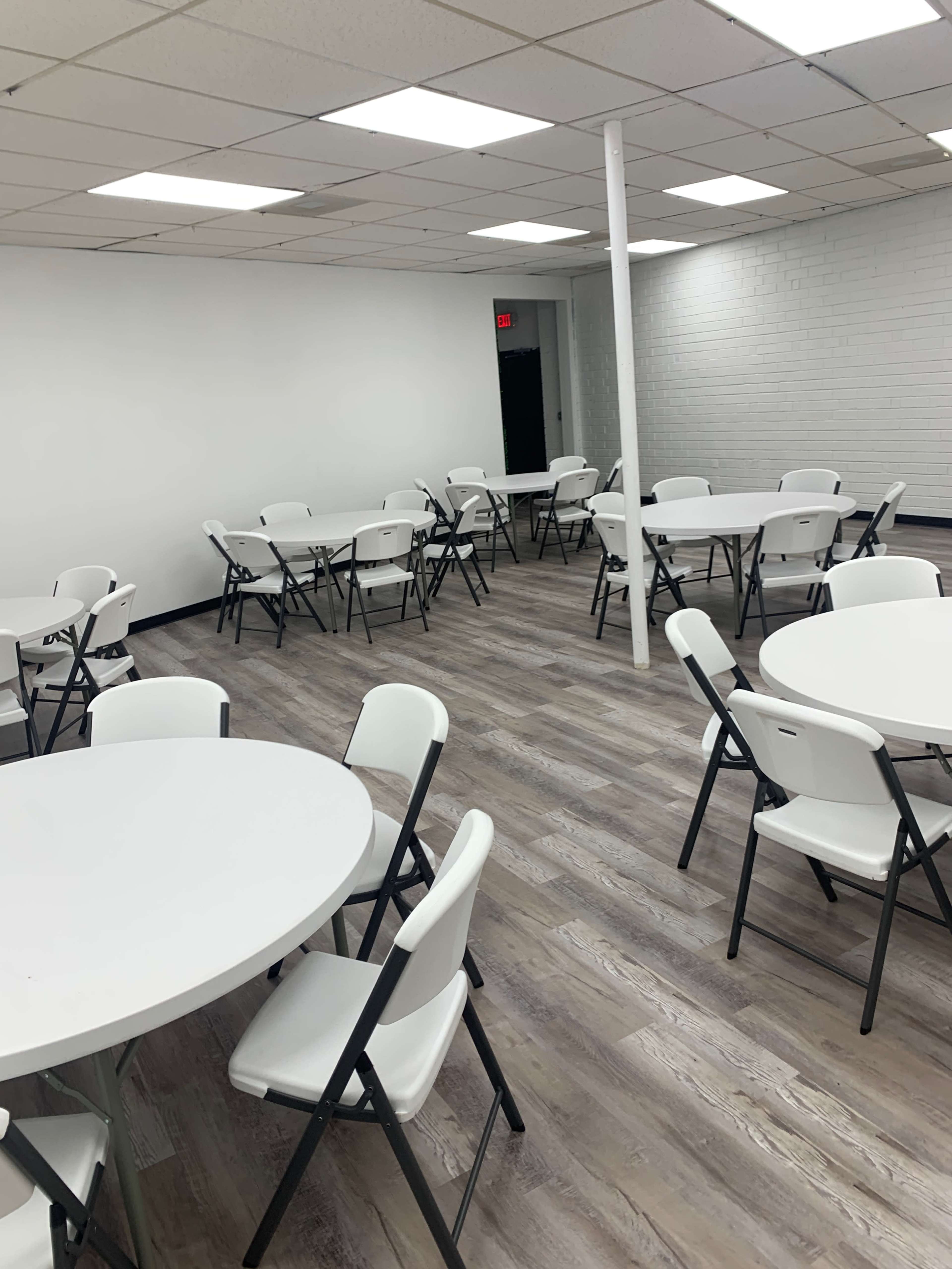 A vacant room features several round tables surrounded by white folding chairs on a wooden floor.