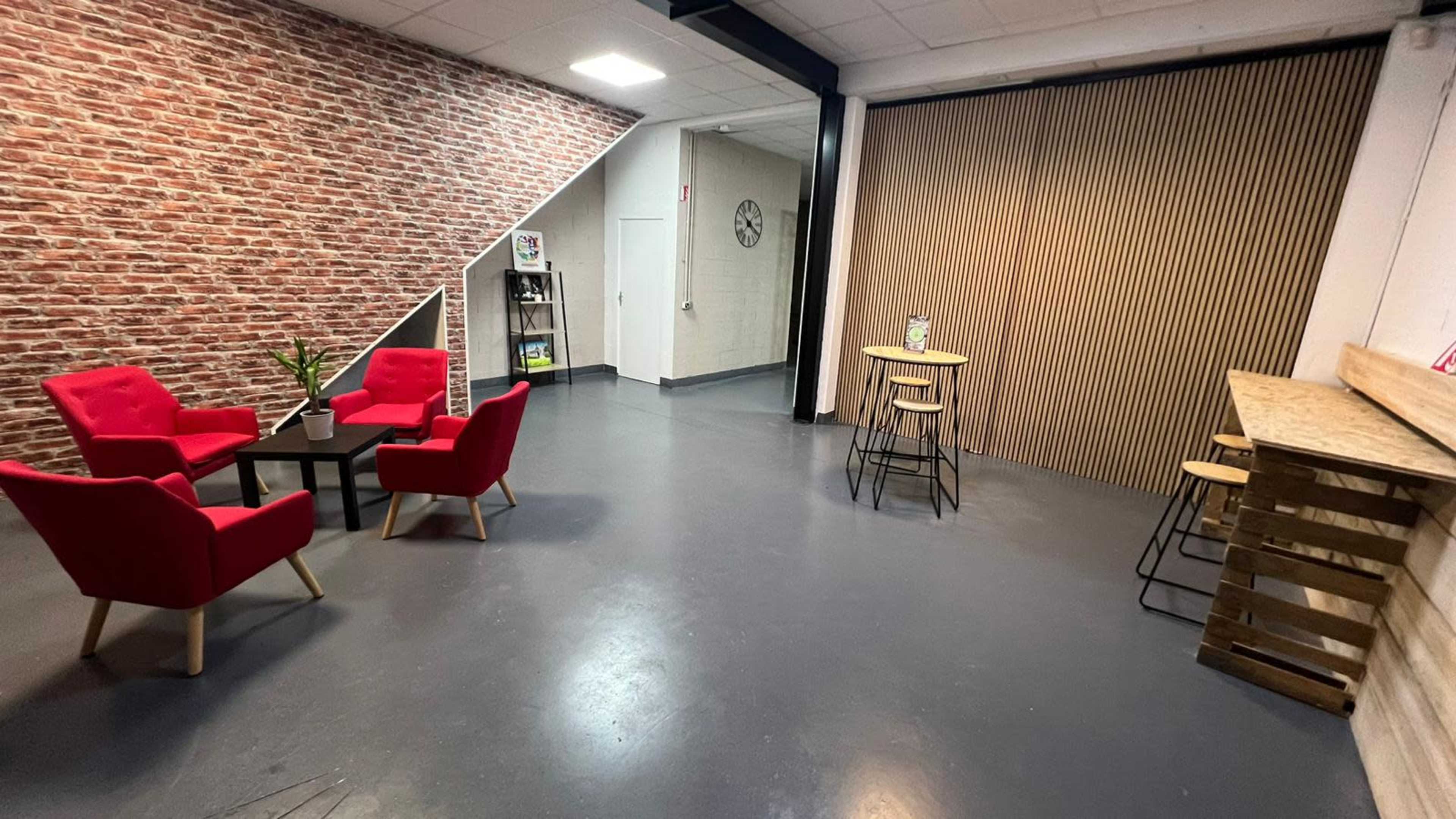 A spacious room with a brick wall, featuring red armchairs grouped around a black table and a wooden counter with bar stools against a backdrop of wooden slats.