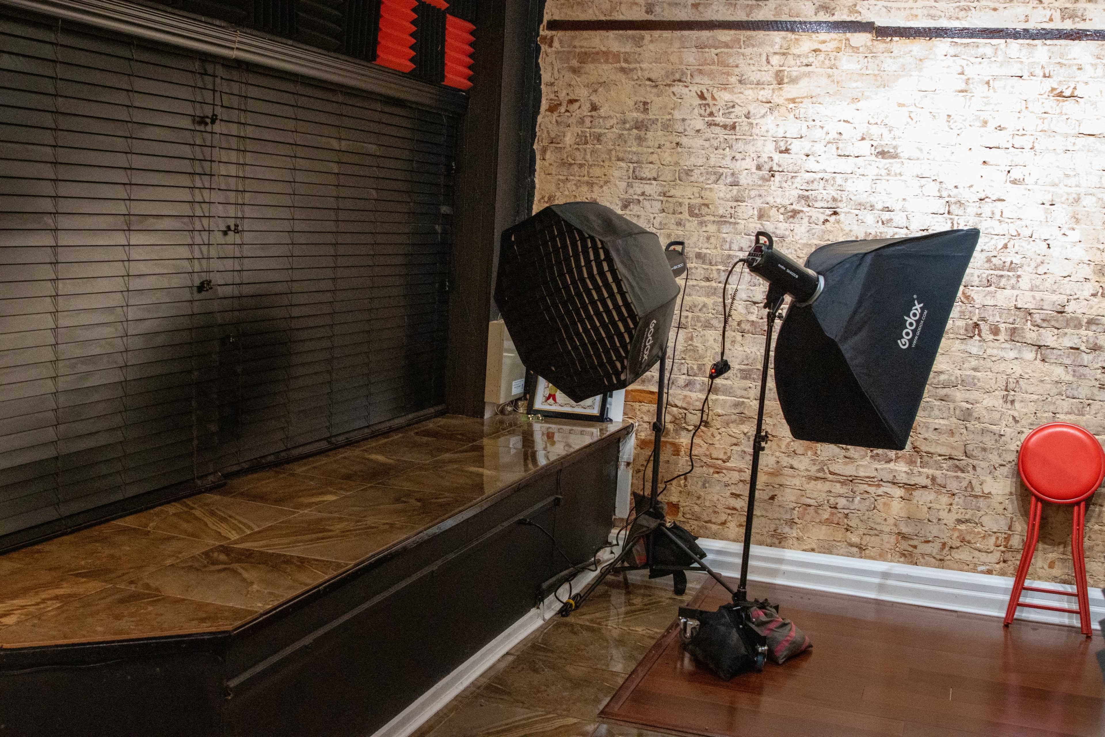 A photography studio setup with two light softboxes directed at a raised platform against a brick wall, accompanied by a red chair in the background.