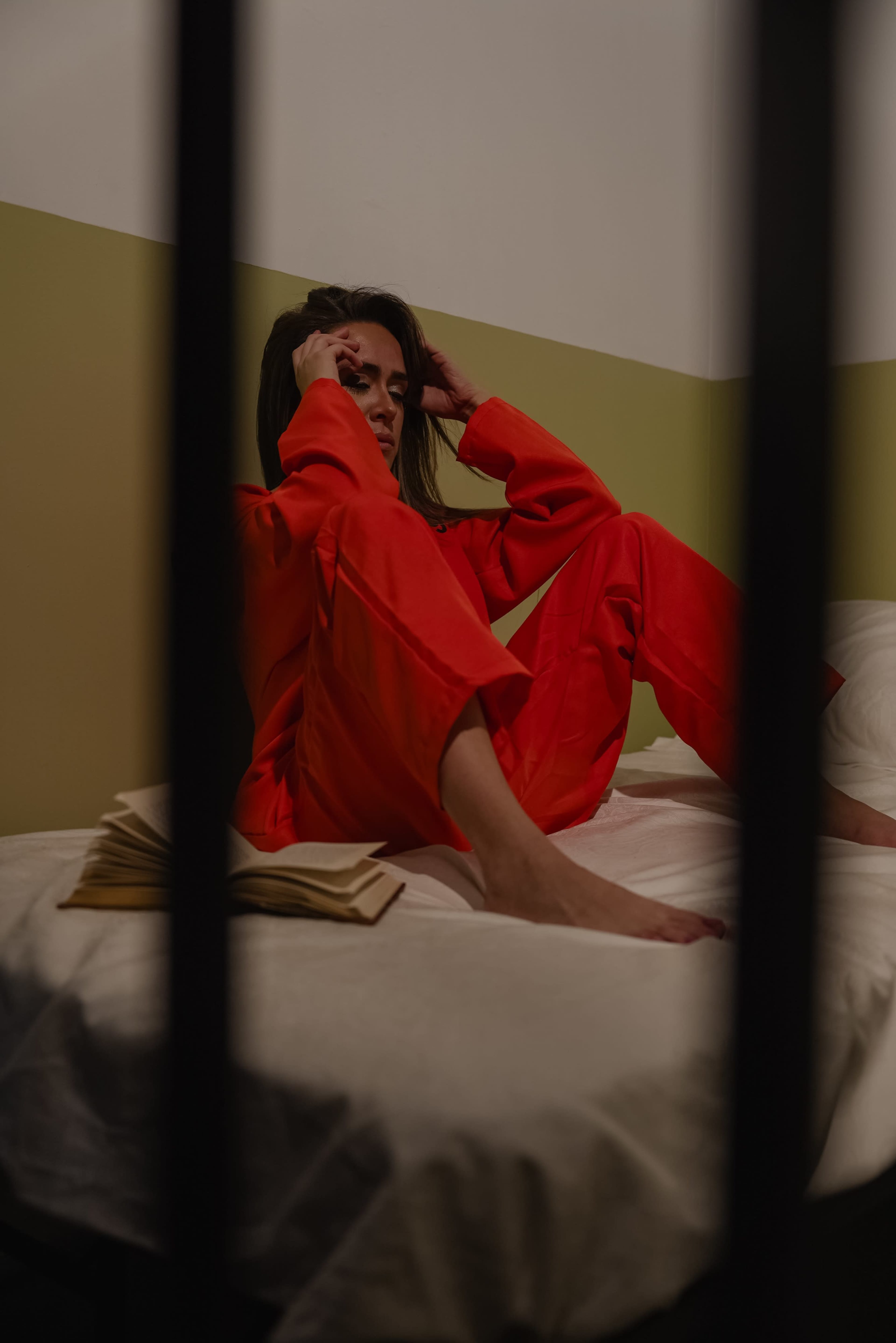 A woman in a red outfit sits on a bed, holding her head with her hands, surrounded by a dimly lit room with muted colors.
