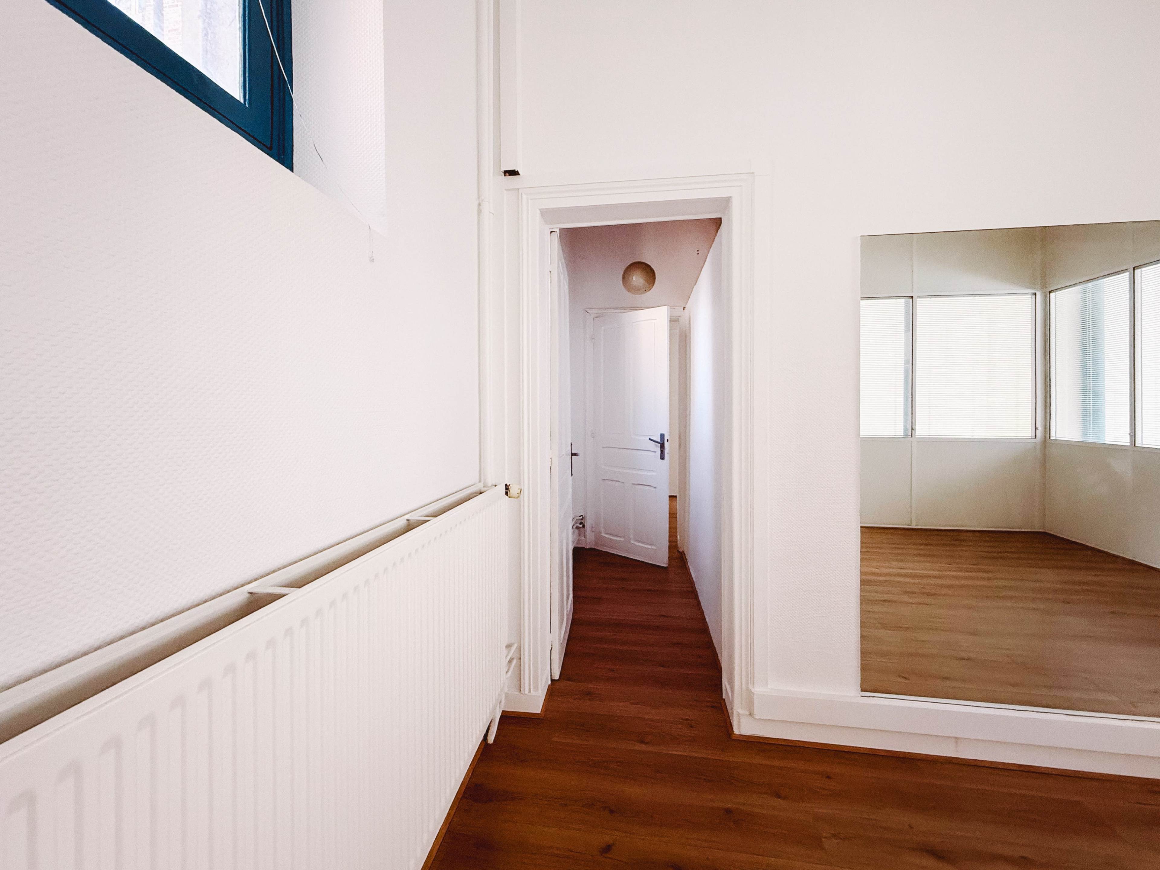 The image shows a hallway leading to a bright room with a large mirror on the wall and a door at the far end.